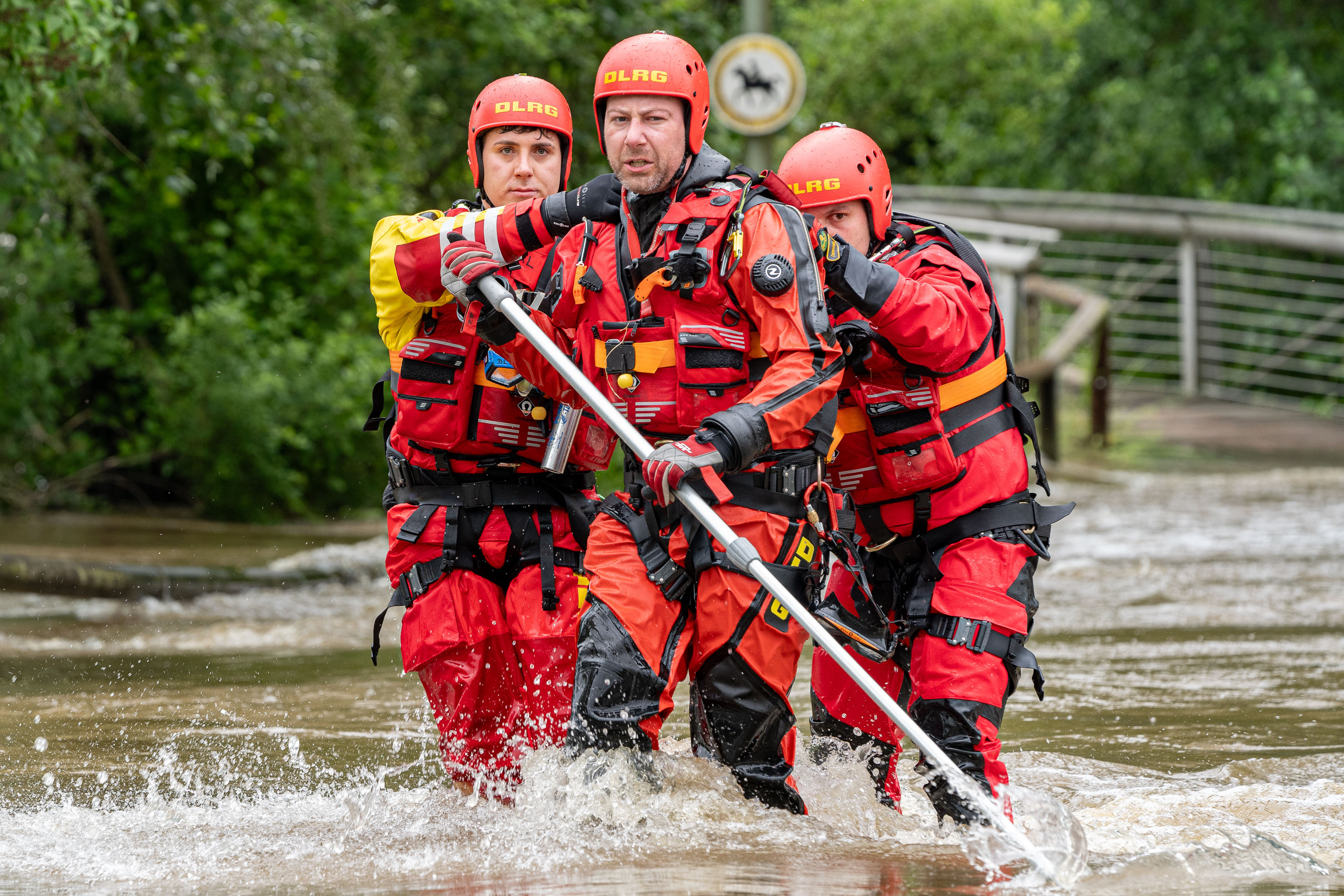 3 Einsatzkräfte der DLRG in roten Schutzanzügen und mit roten Helmen versuchen sich einen Weg durch das Hochwasser zu bahnen. Der vorderste Helfer hält eine Stange in der Hand, um nicht den Halt zu verlieren. die restlichen beiden halten sich an ihm fest.