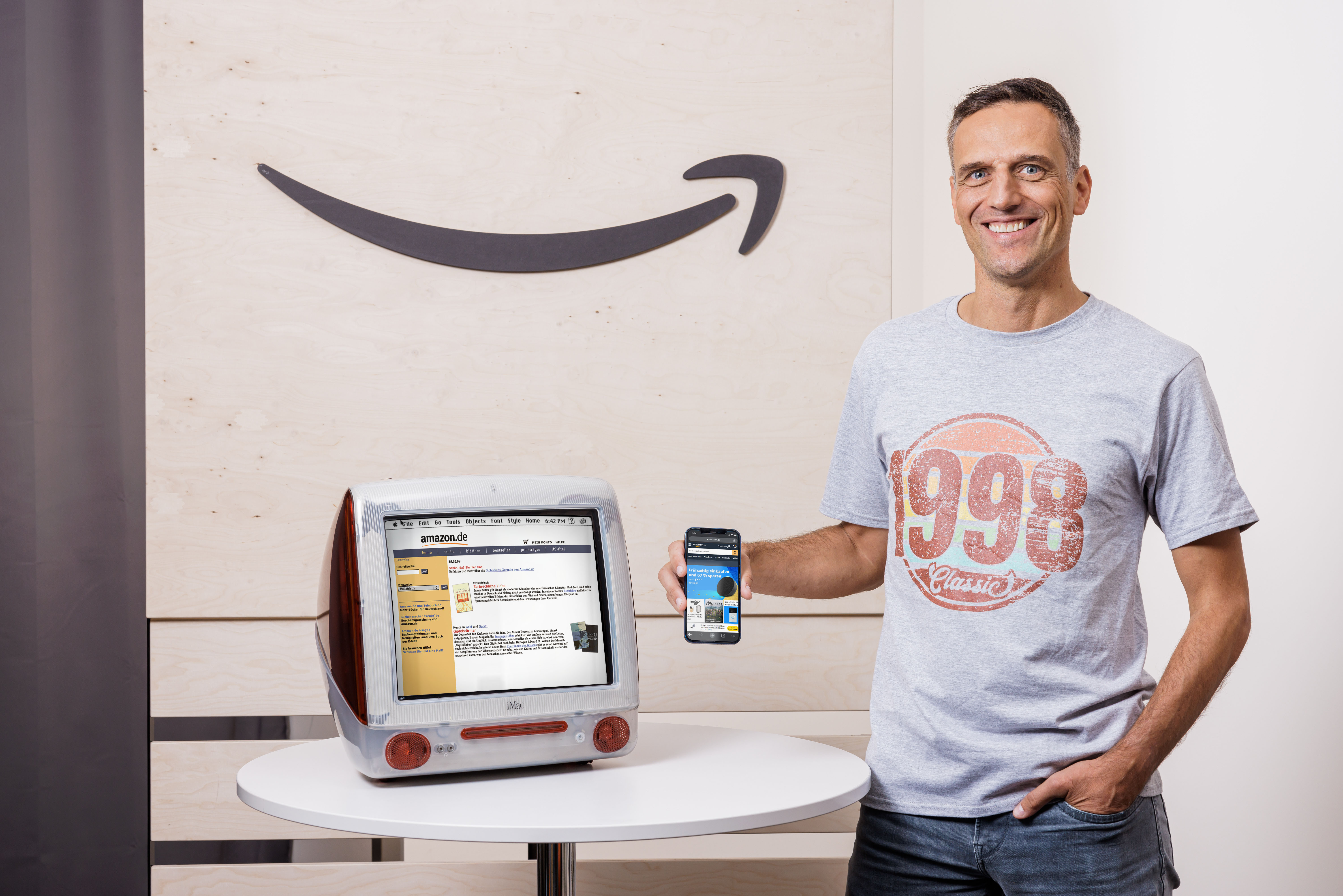 Amazon Deutschland gibt es seit 25 Jahren. Rocco Bräuniger hält ein Smartphone mit der Amazon App vor einen PC mit der Amazon Website.