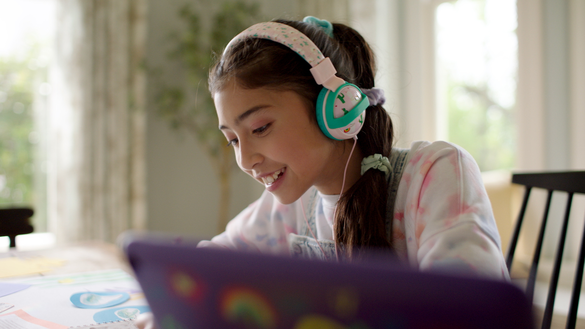 A pre-teen girl wearing headphones looks down at something in front of her, on a table. There is a tablet device in the foreground.