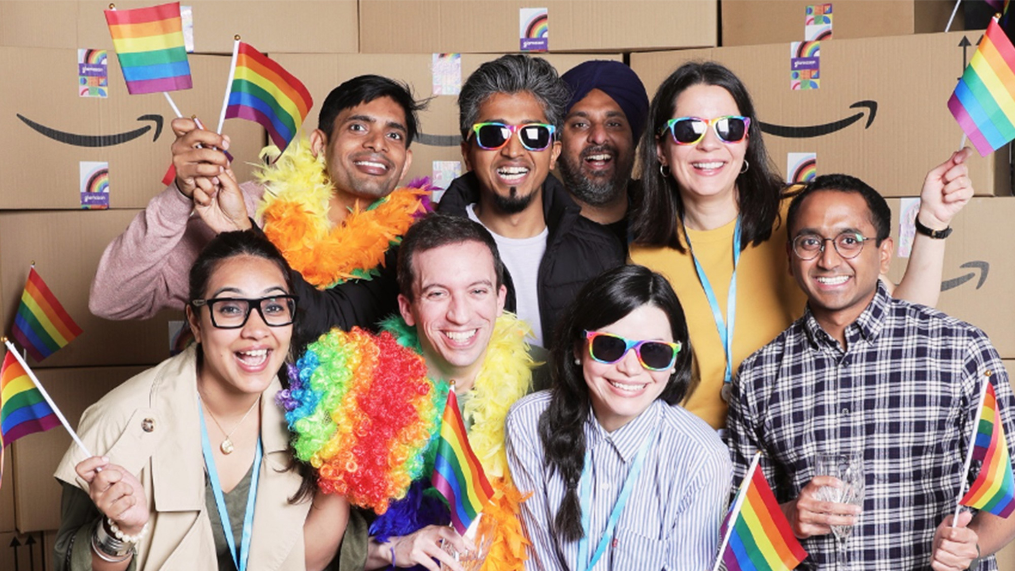 A group image of Amazon employees celebrating Pride and waving the LGBTQIA+ flag. There are boxes with the Amazon logo in the background.