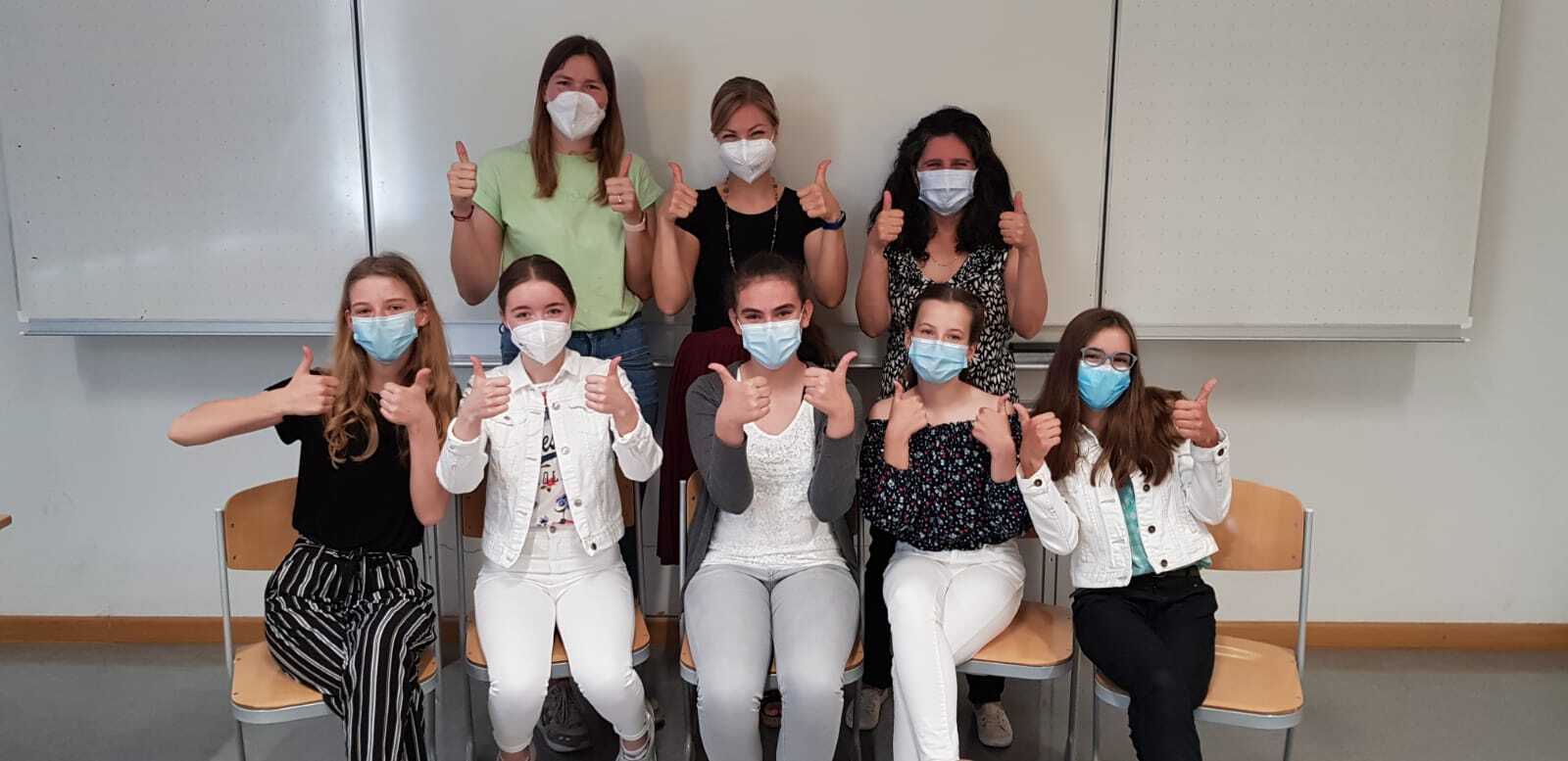 Mädchen mit Masken sitzen auf Stühlen und schauen in die Kamera.