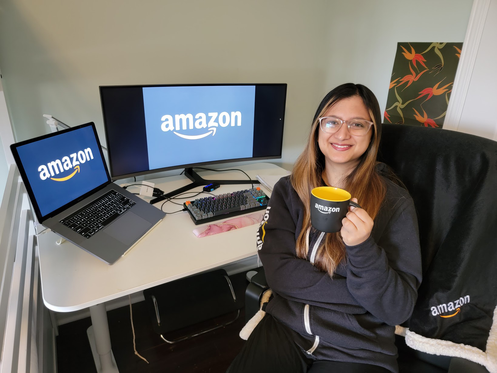 An image of a woman smiling for a photo at her desk. Behind her on the desk are two monitors with the Amazon logo on them. She is wearing an Amazon sweatshirt and holding an Amazon mug.