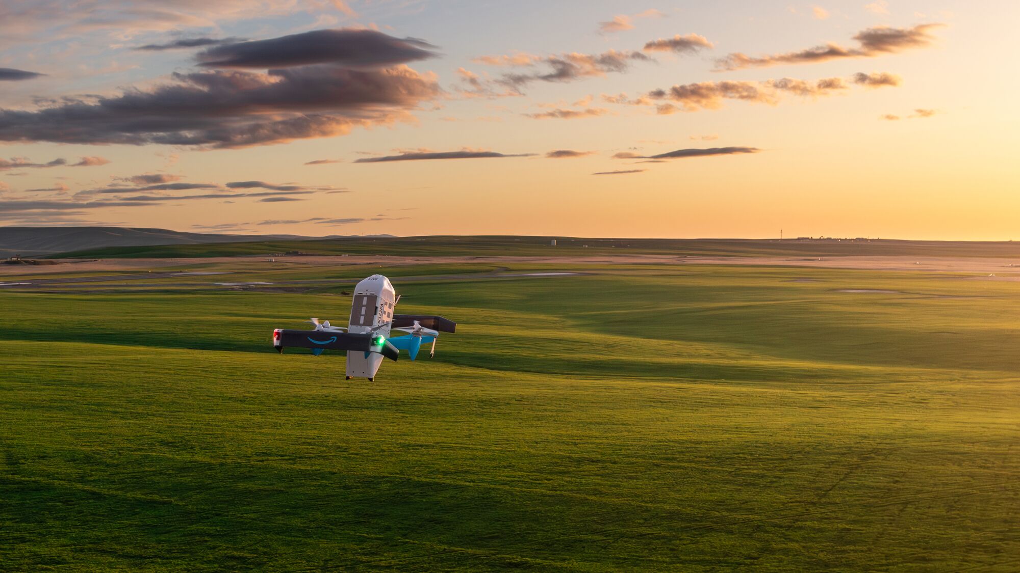 An Amazon delivery drone hovering low over a vast green grassland at sunset