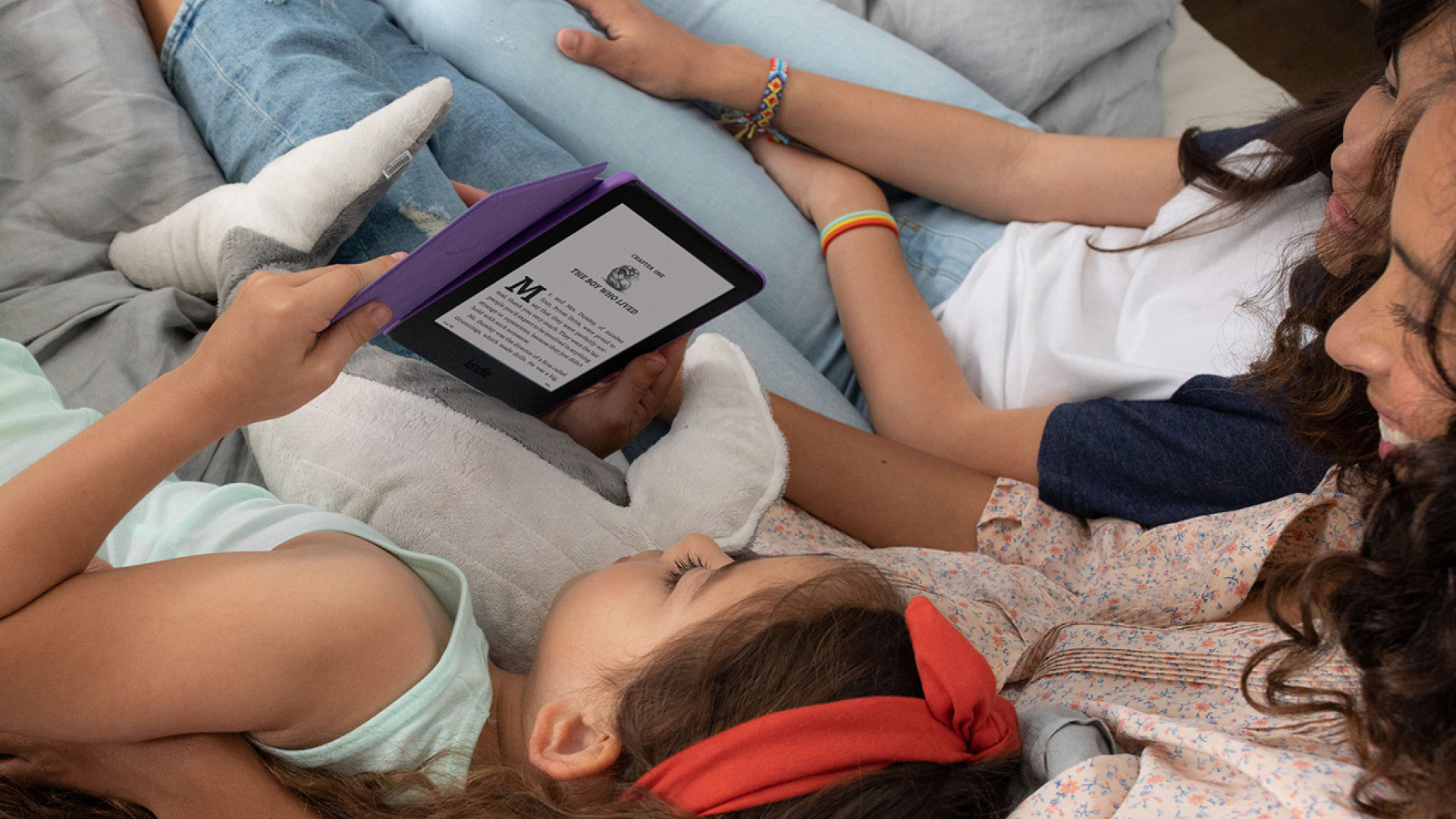 An image of three girls reading on a Kindle device while laying down. The girl holding the device has a stuffed animal next to her and is wearing a red headband. 