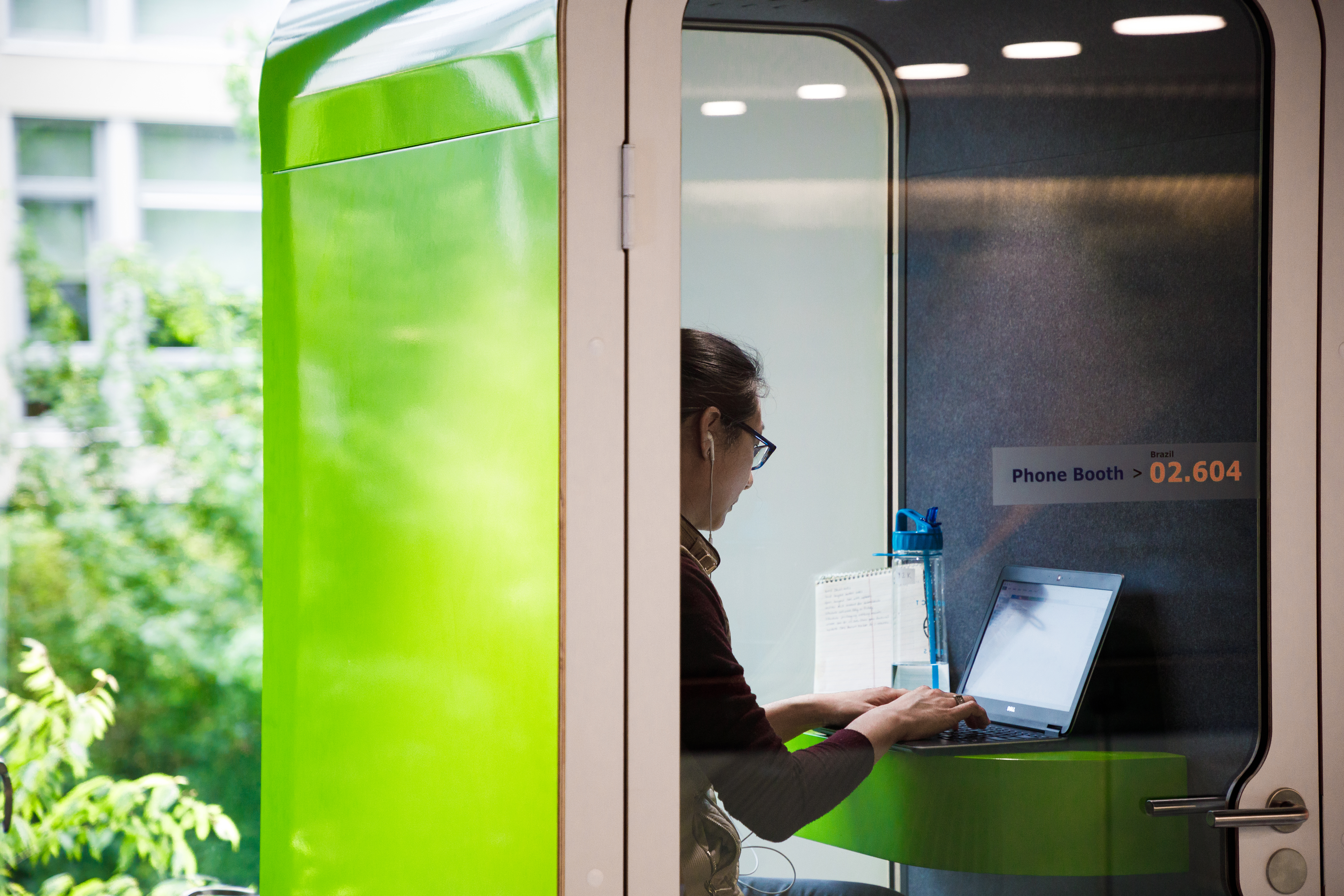 A woman wearing Apple earbuds is typing on a computer inside of a green, private phone booth at Amazon's HQ in Seattle, WA. She has a water bottle sitting on her desk. 