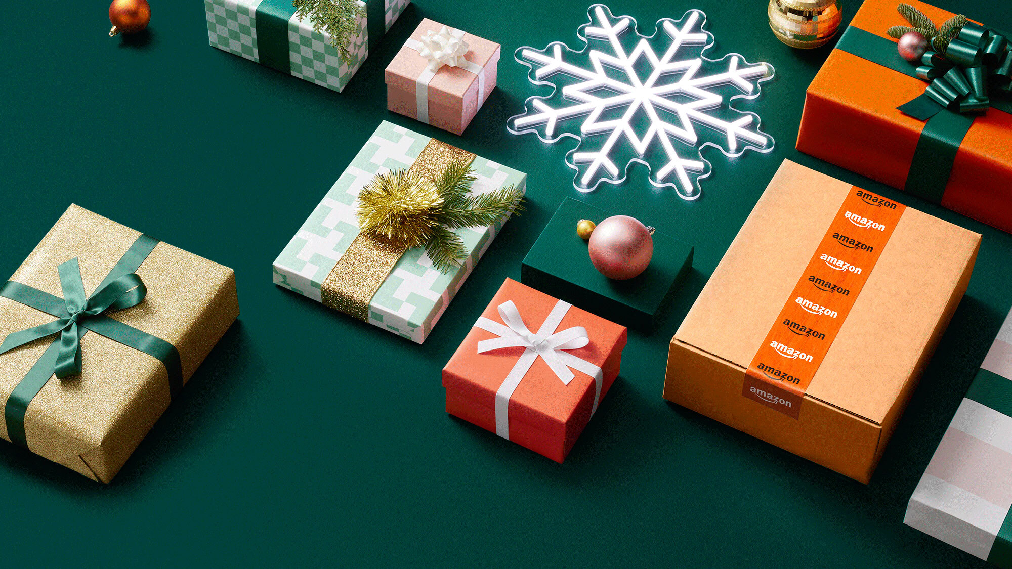 an image with a dark green background. There are various wrapped gifts in colorful packaging with ribbons, an Amazon box, and a light-up snowflake.