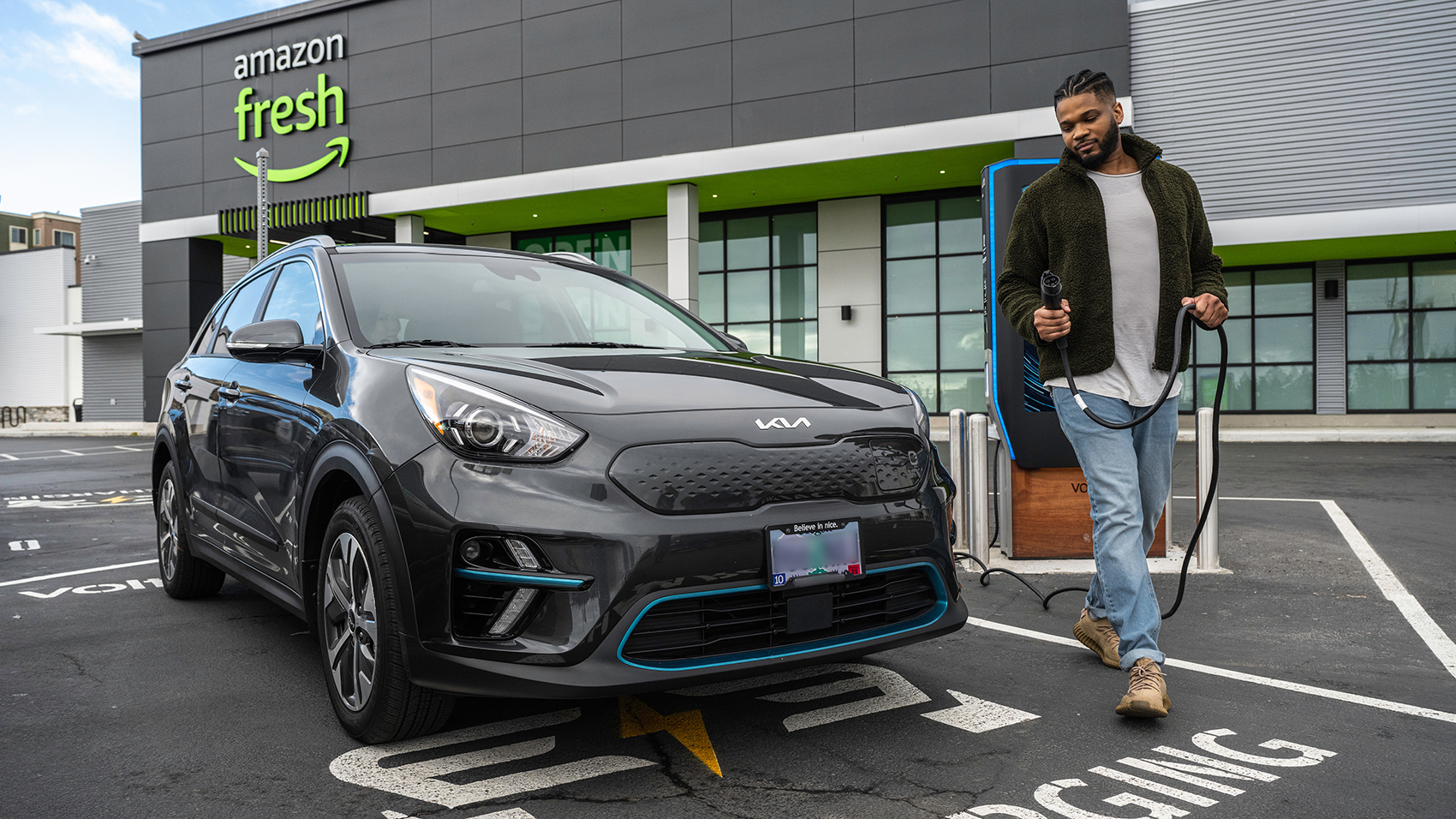 An image of a man walking around an electric car with a cord that he is going to plug in to charge it. The car and the man are in the parking lot in front of an Amazon Fresh store.