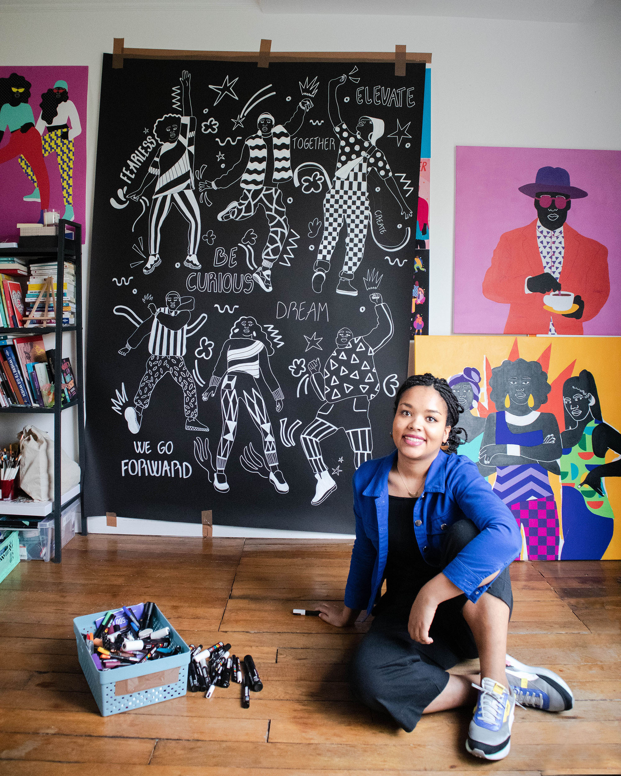 a Black woman, Aurelia Durand, sits on the wooden floor of her studio. Her artwork is displayed on the wall behind her, with saturated colors, and stylized people. 