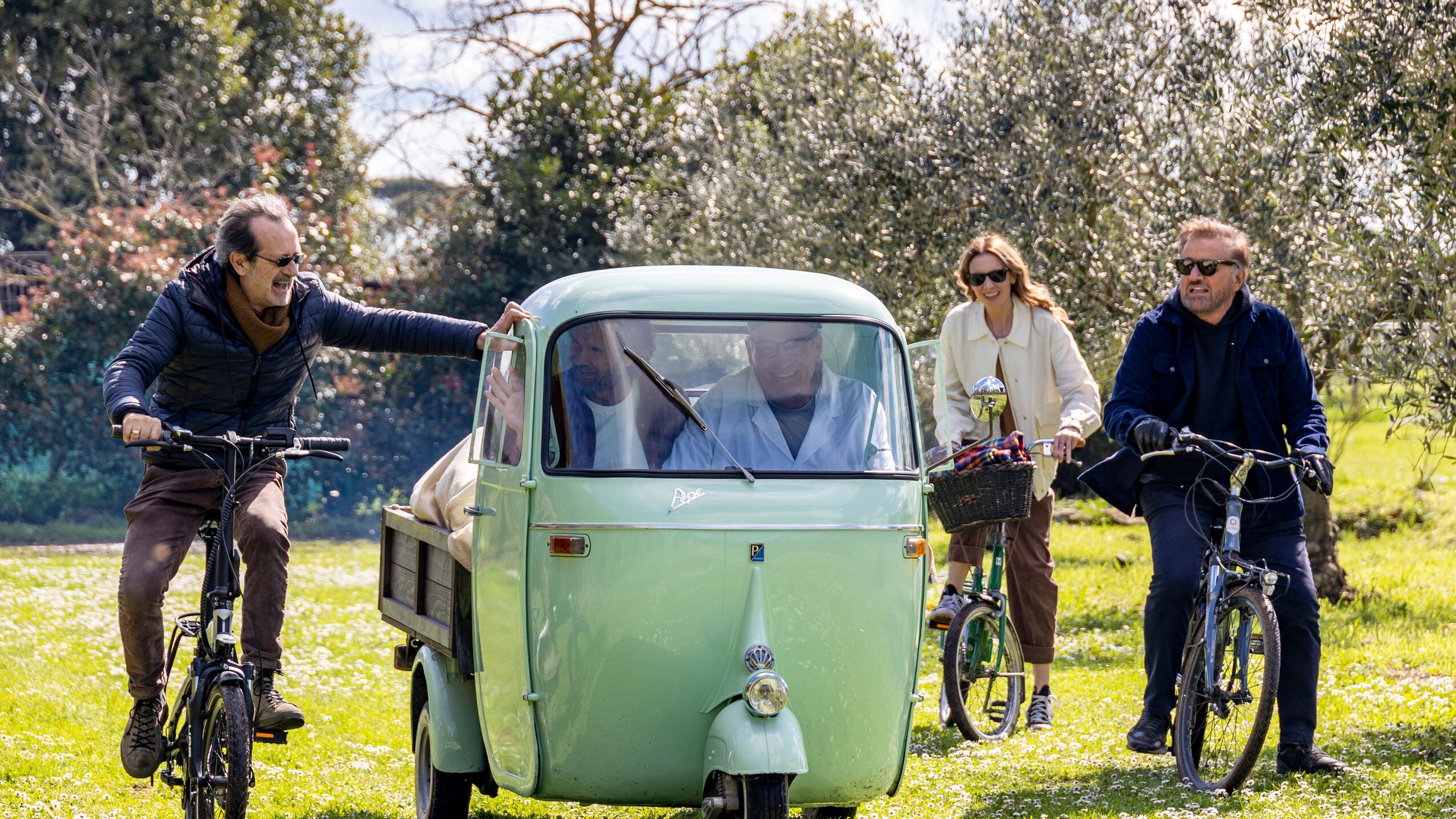 Vintage Piaggio Ape and cyclists gather on grassy field surrounded by trees while filming Dinner Club