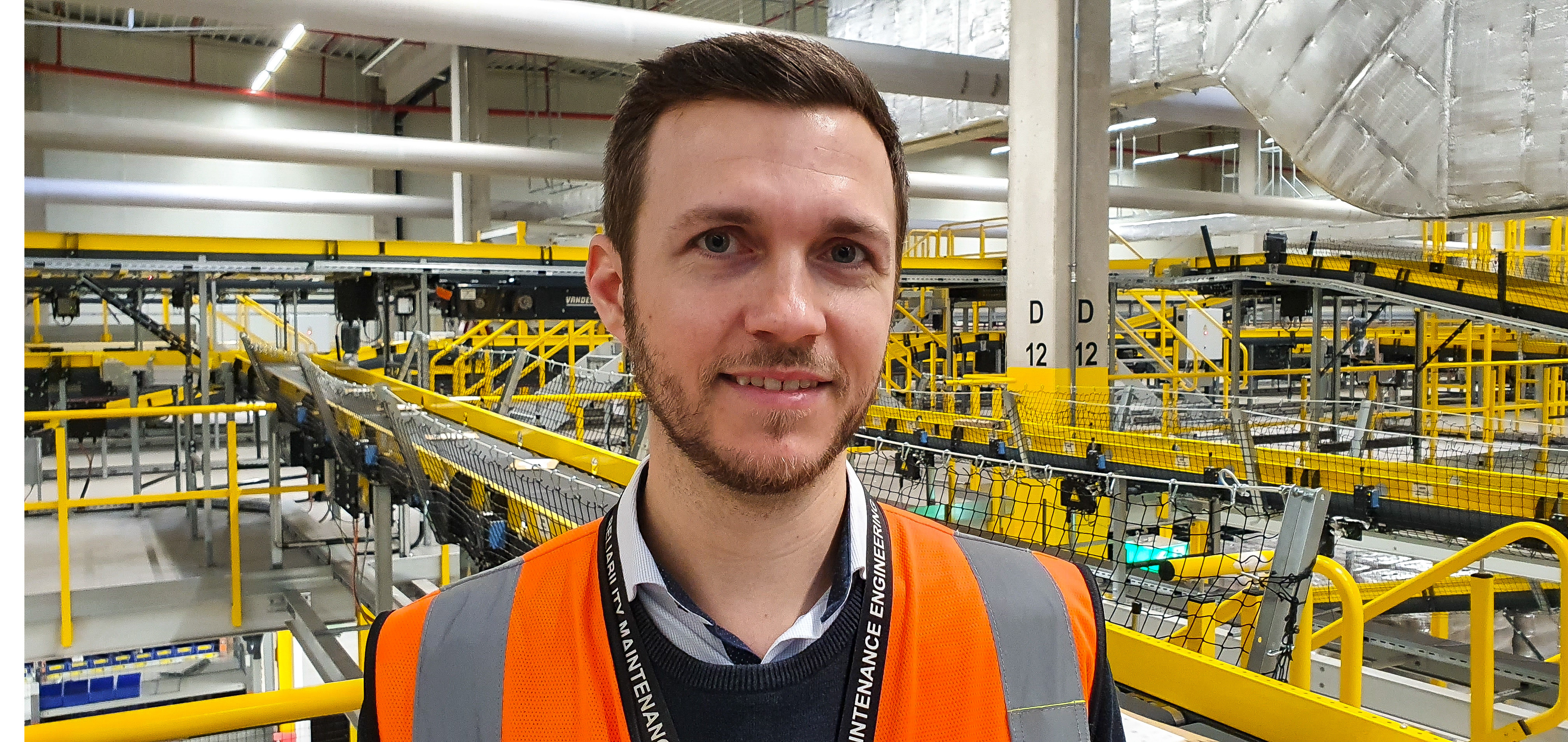 Ein junger Mann mit braunen Haaren und Bart vor einer Förderanlage in einem Logistikzentrum.