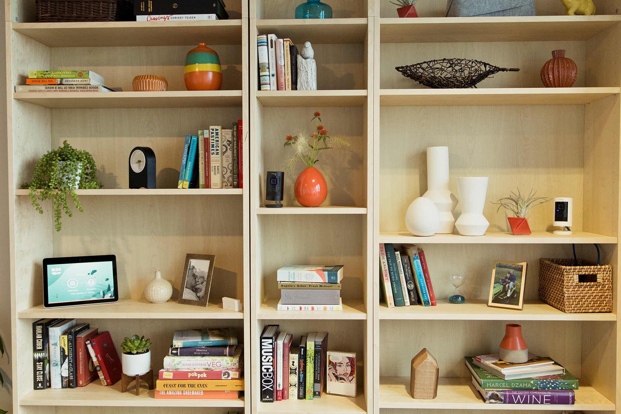 A bookshelf with books, decor, vases and baskets, clock, Echo Show, and Ring camera, illustrating a Smart Home setting.