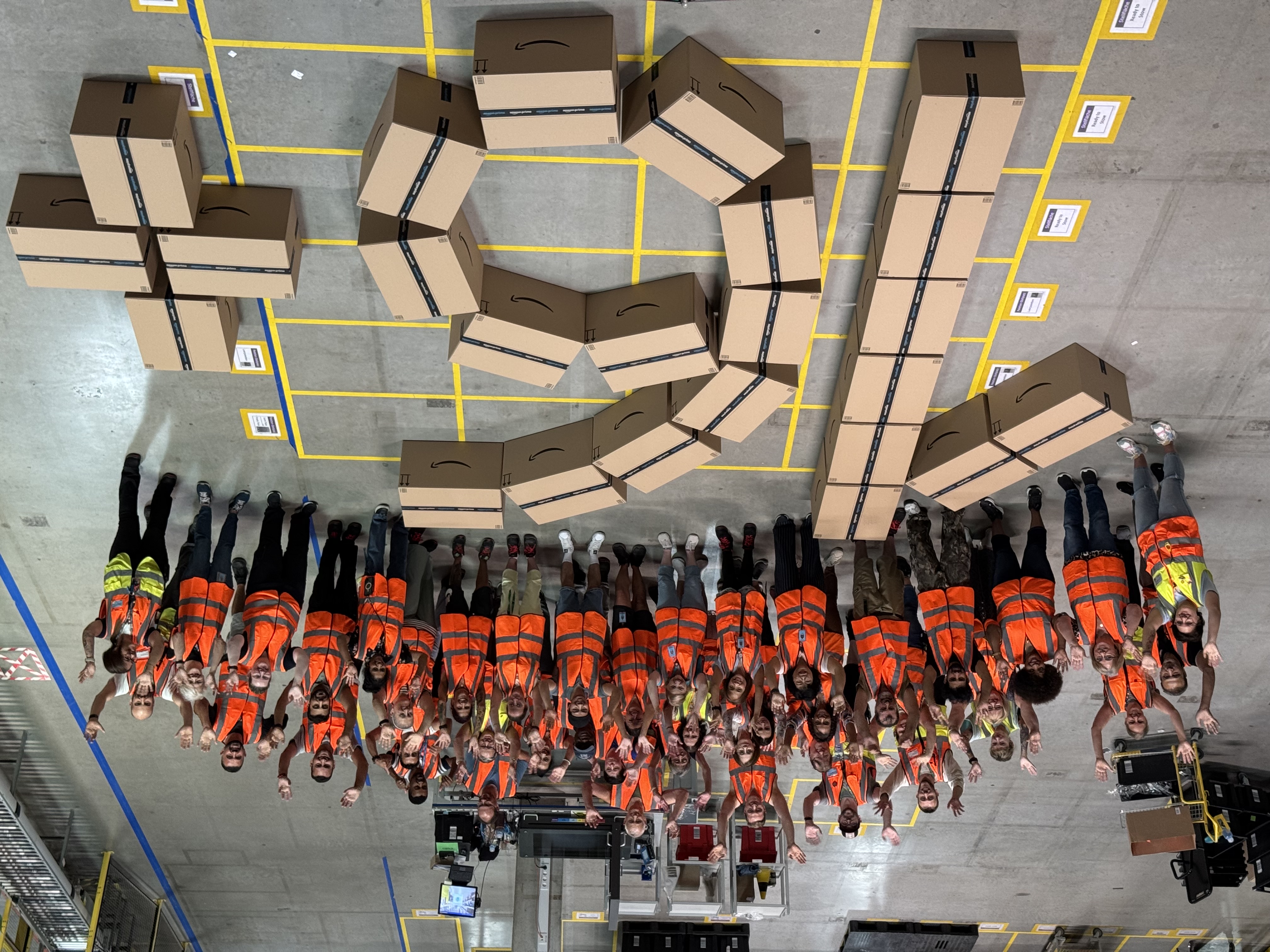 Mitarbeitende stehen im Logistikzentrum um die aus Paketen gelegte Zahl 16+