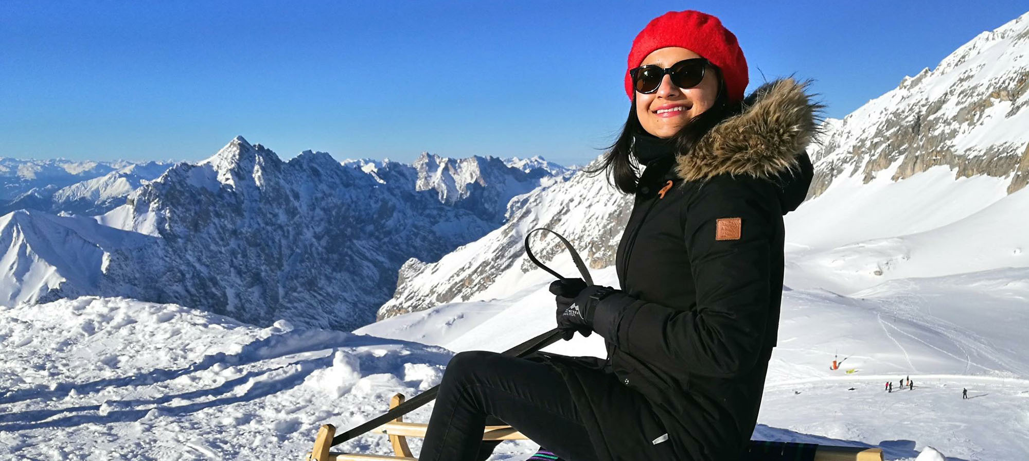 Mehak in den Alpen beim Schlitten fahren. Sie trägt eine rote Pudelmütze und eine Sonnenbrille. Im Hintergrund sieht man die schneebedeckten Alpen.