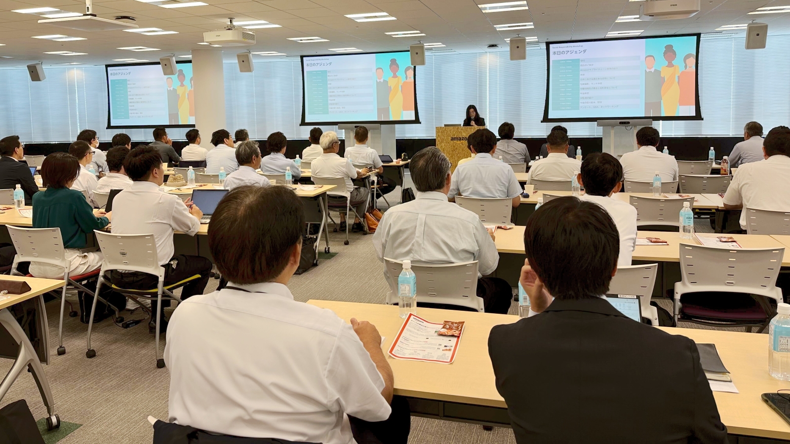 presentation in modern conference room with attendees seated at individual desks facing three projection screens