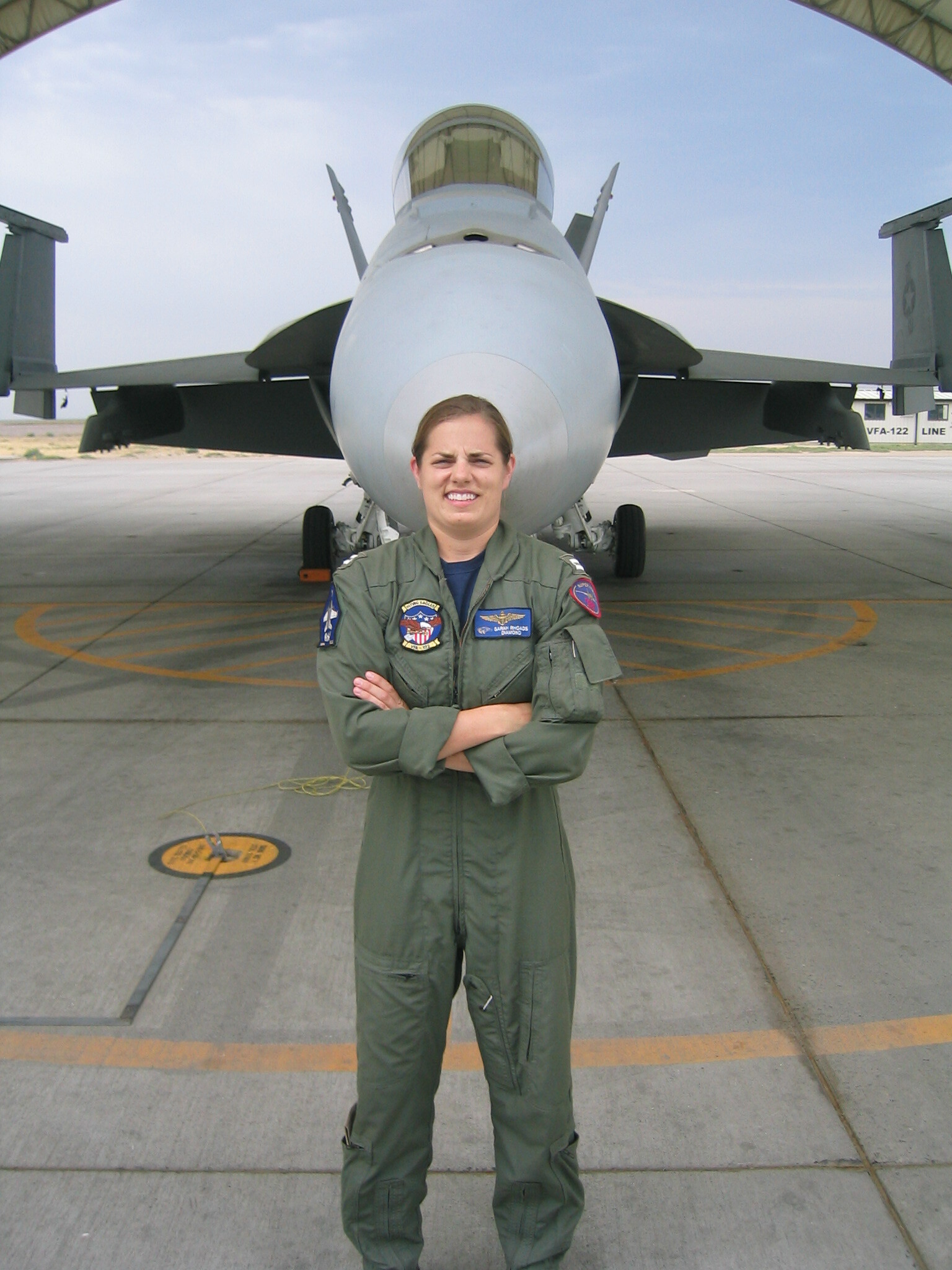 A woman in a United States Navy flight suit stands in front of a fighter jet with her arms crossed. 