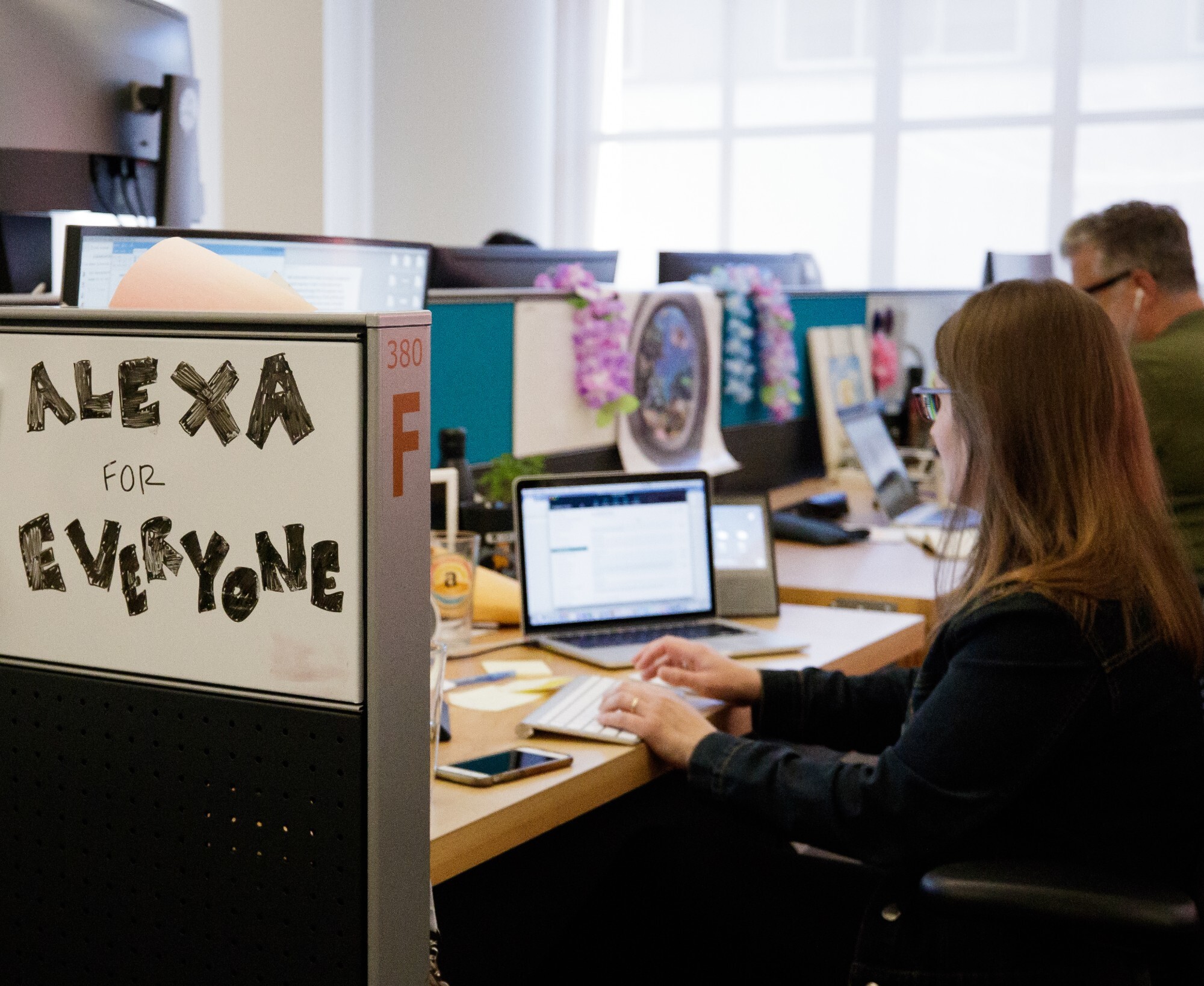 Two people sit at their desks which are side-by-side. There is a white board that reads "Alexa for everyone"