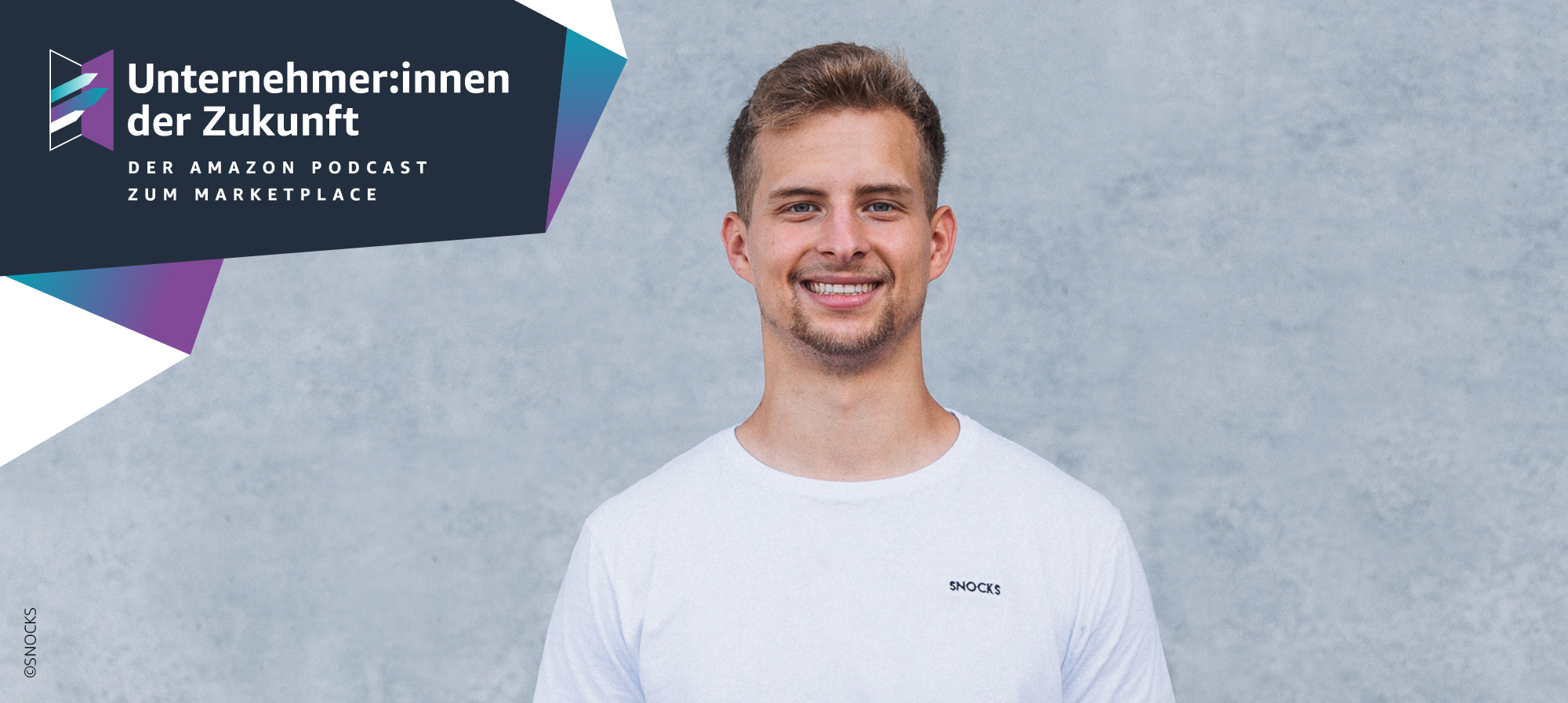 Ein Mann mit blonden Haaren und einem weißen T-shirt steht vor einer grauen Wand und lacht in die Kamera.
