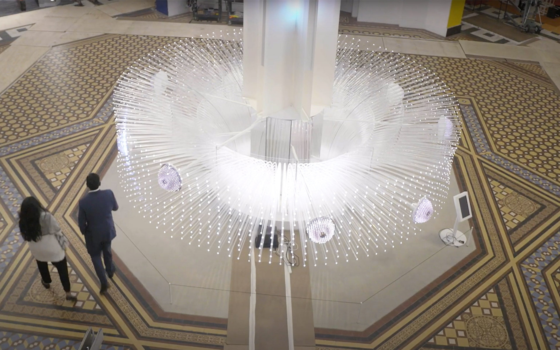 An image of a large white structure with multiple glass shoots coming out of it. At the end of each shoot is a small light. There are small circles within the structure that have smaller clusters of lights with a purple tint. The structure stands in the middle of a museum, and two people are walking by it.