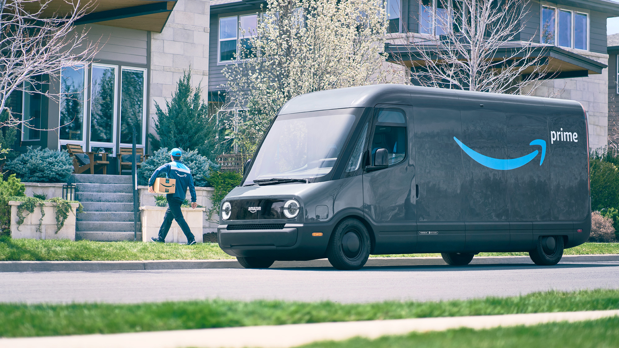 An Amazon delivery driver walks a package to a customer's front door while the Rivian delivery vehicle is parked on the street.