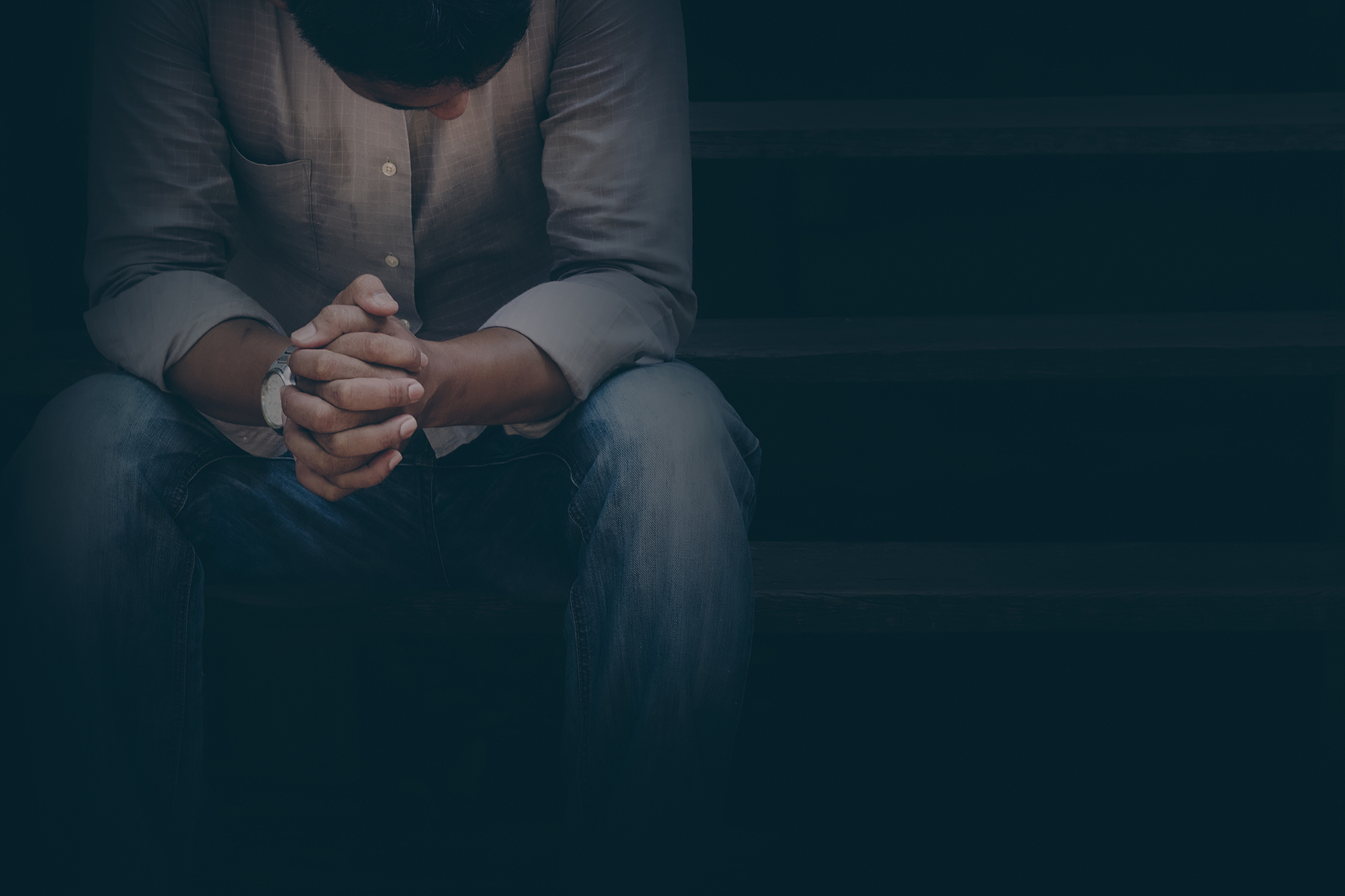 A man in a button-down and jeans sits with his hands folded, looking downward in a dark setting.