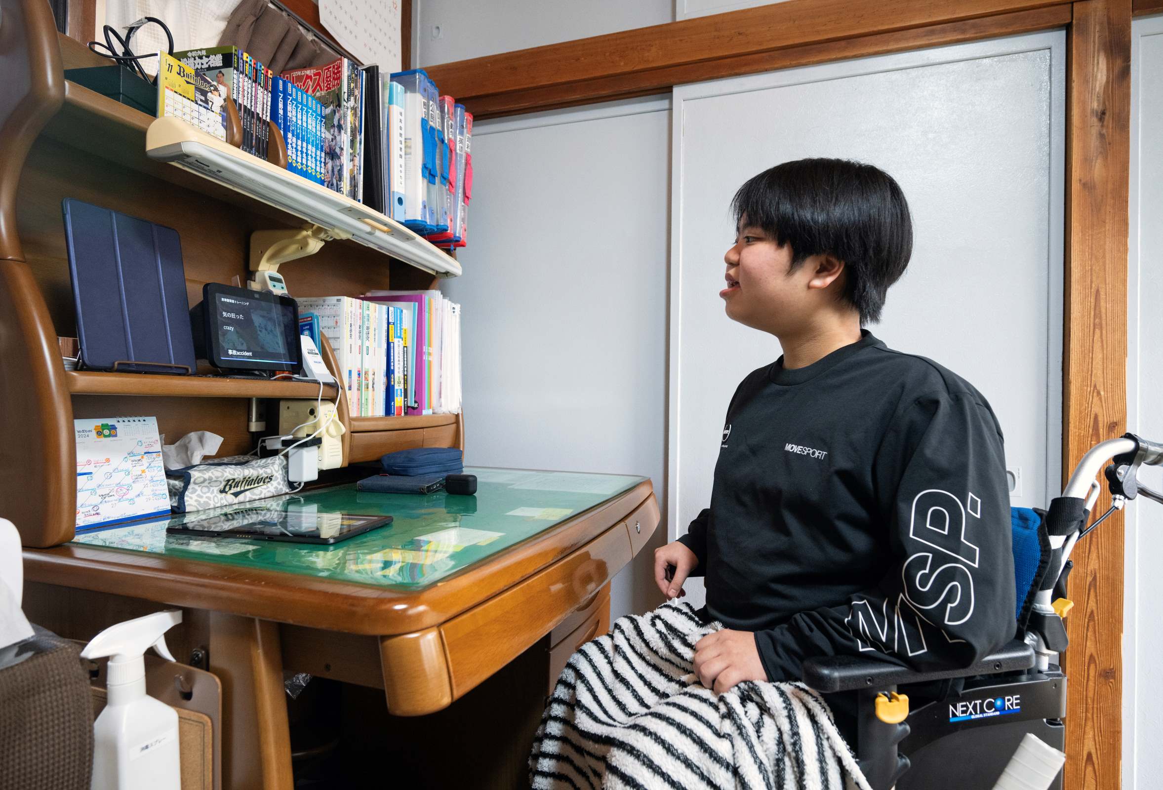 車椅子に座っている男性がデスクの上に置かれたタブレット(Echo Show 8)を見ている。タブレットの他には様々な本やホルダーなどがきちんと並んでいる。