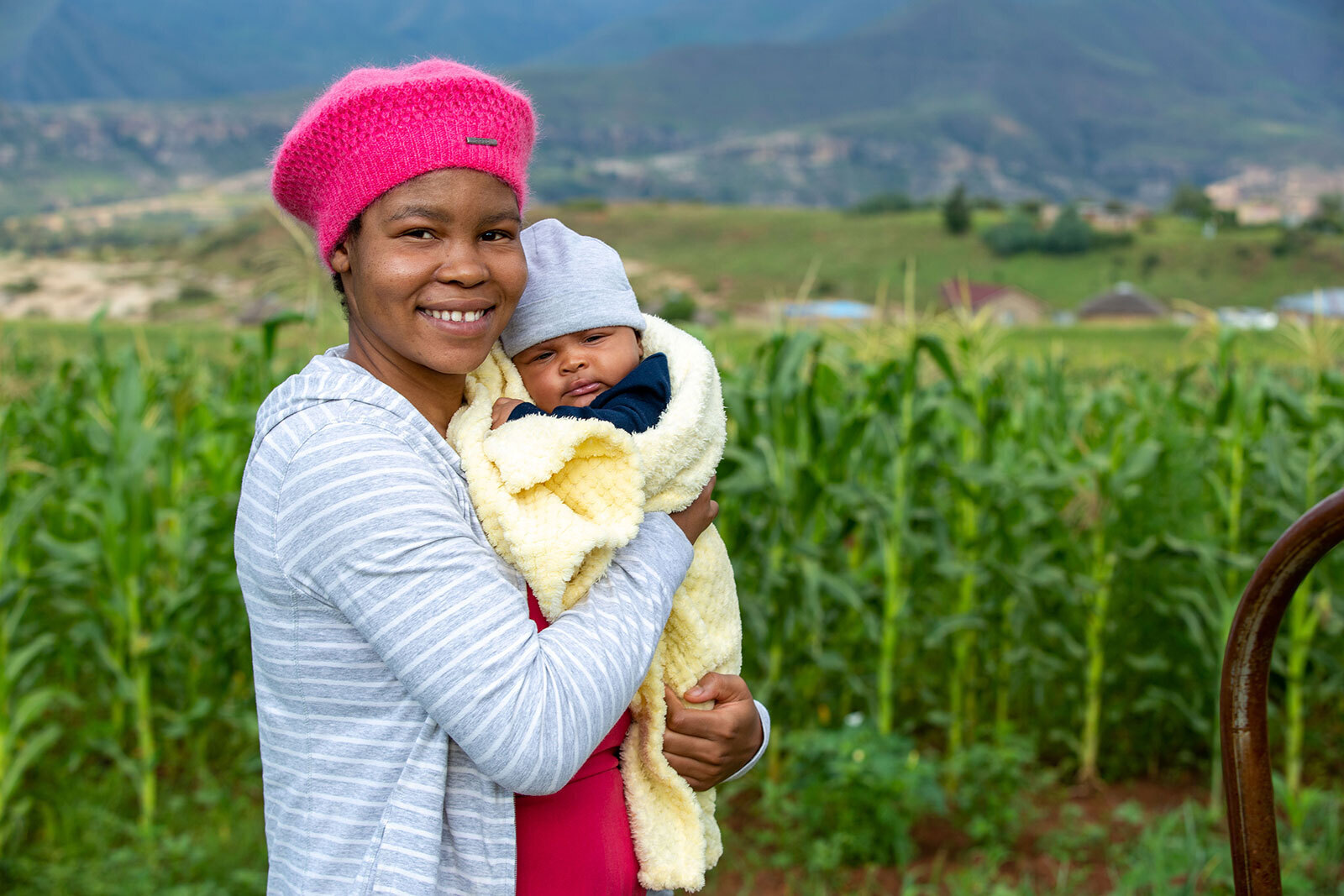 An image of a woman smiling for a photo while standing in front of a green field. She is holding a newborn baby wrapped in a light yellow blanket.