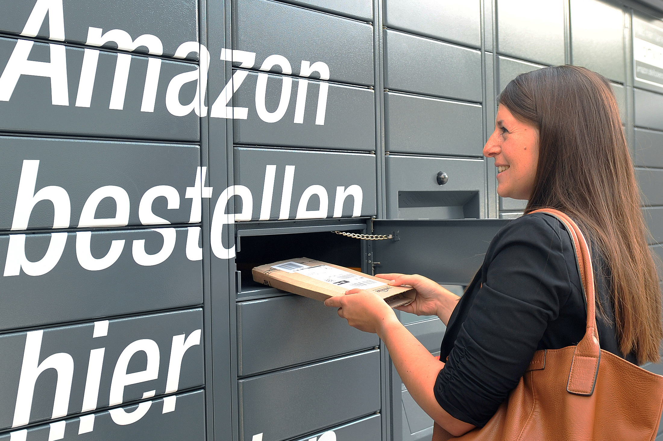 Der Bildausschnitt zeigt graue Paketschließfächer und den weißen Schriftzug "Amazon bestellen hier ...". Rechts ist eine junge Frau mit langen braunen Haaren zu sehen, die ein längliches, flaches Amazon Paket aus dem Schließfach entnimmt.