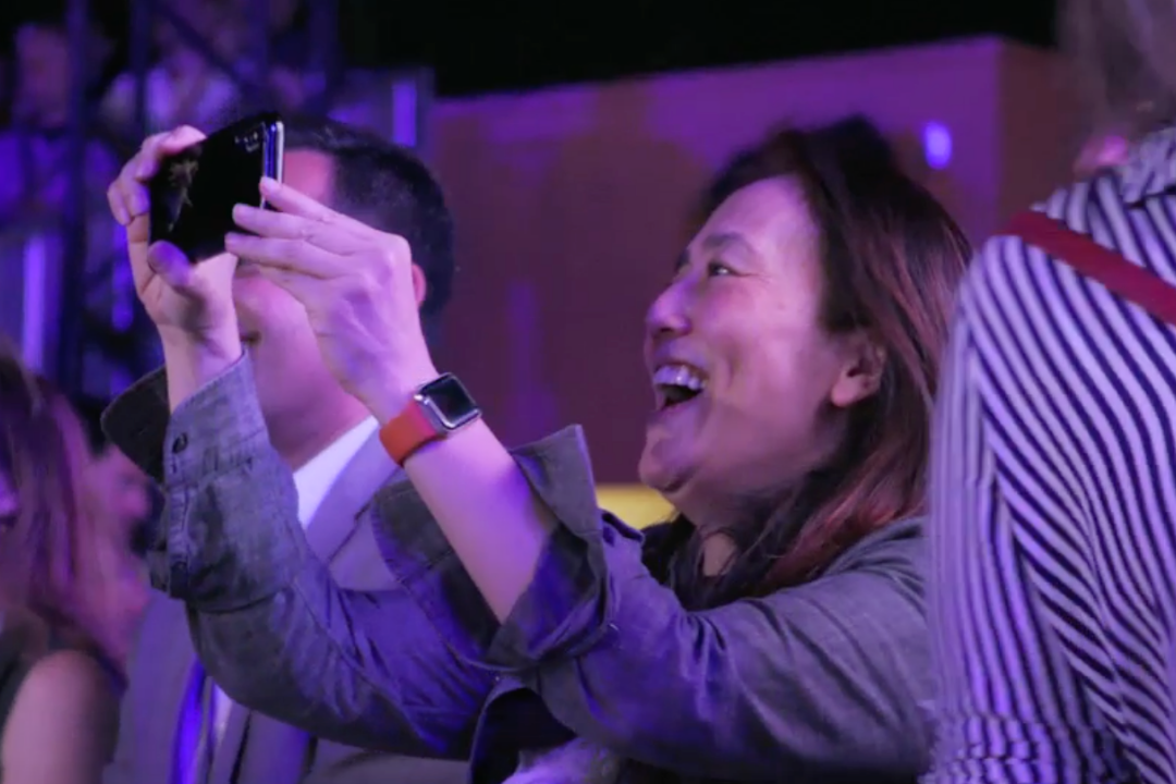 An image of a women smiling while filming something on stage.