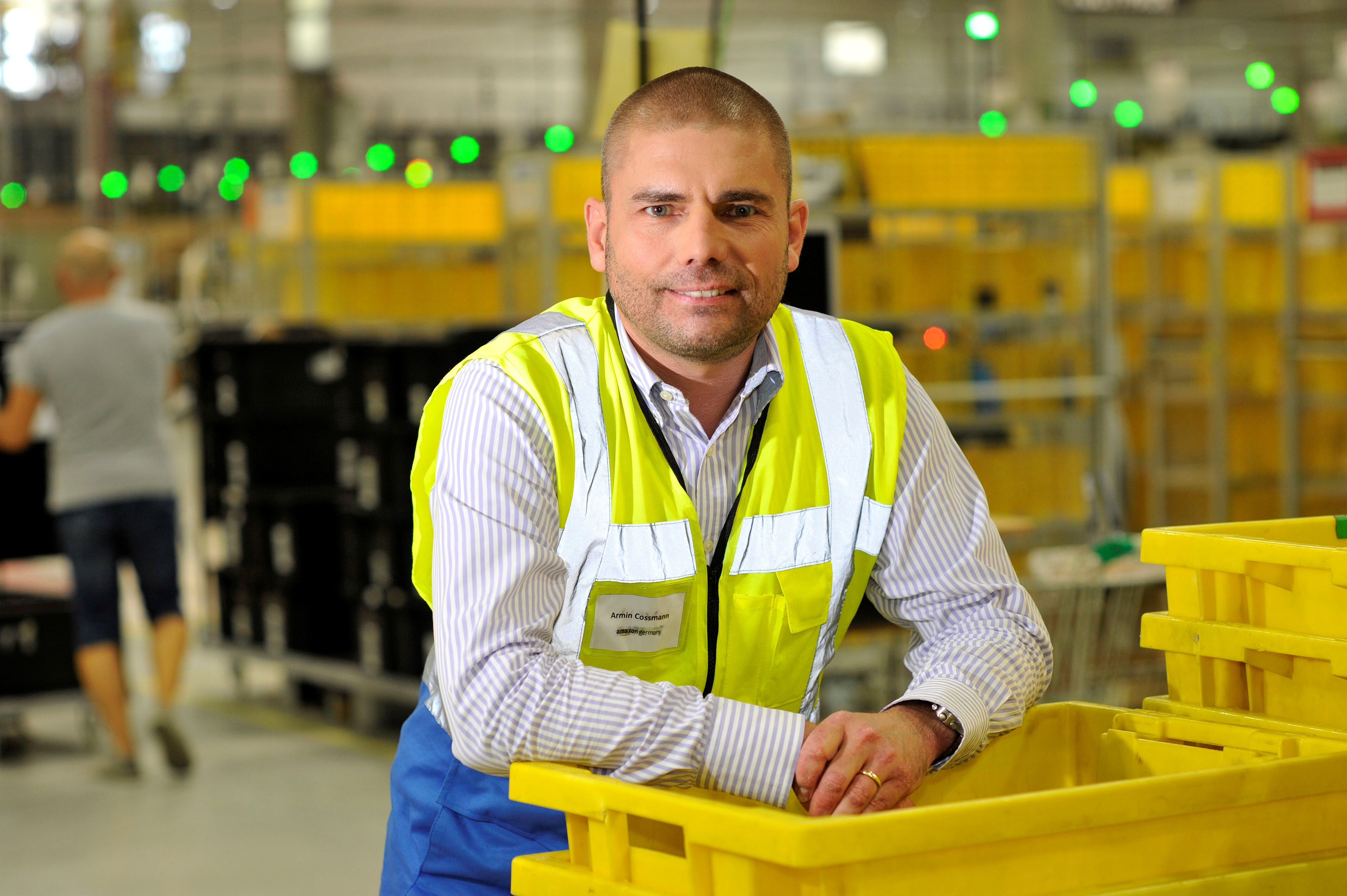 Ein Mann in gelber Sicherheitsweste lehnt sich lässig auf einem Stapler gelber Transportbehälter im Logistikzentrum