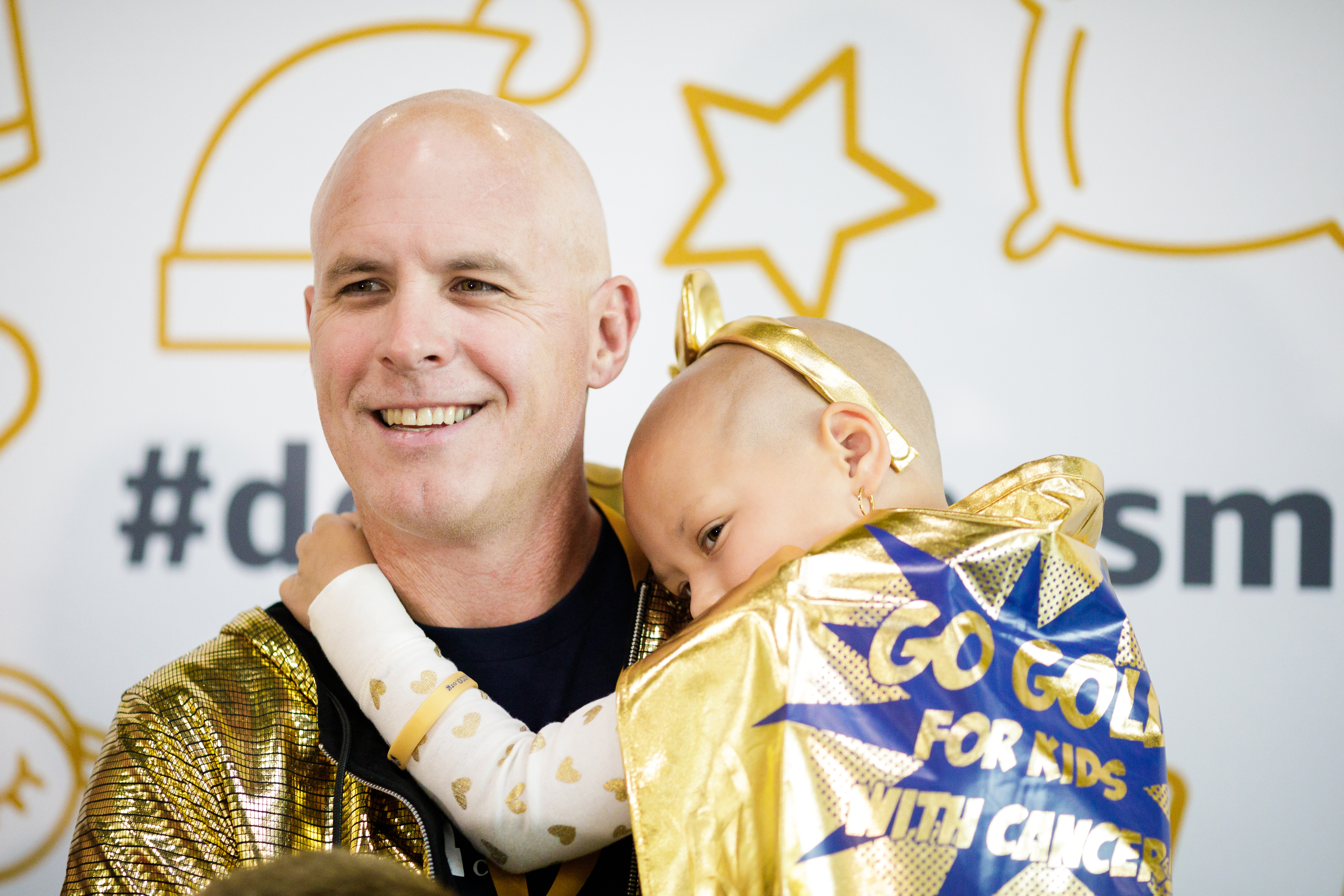 Ein Mann hält ein Kind im Arm: Beide sind haarlos und tragen goldene Umhänge. Das Kind ist von hinten zu sehen. Auf dem Umhang steht: Go Gold For Kids With Cancer.