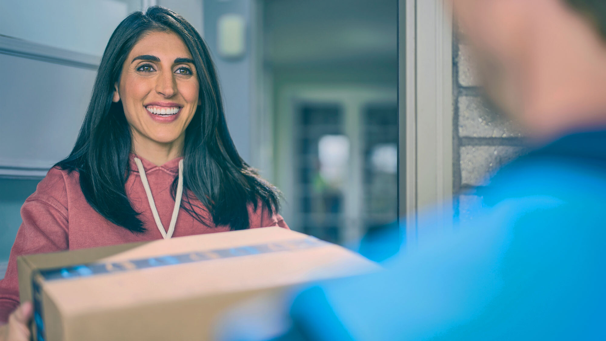 A woman with long dark brown hair smiles as she accept her Amazon package from a delivery driver. 