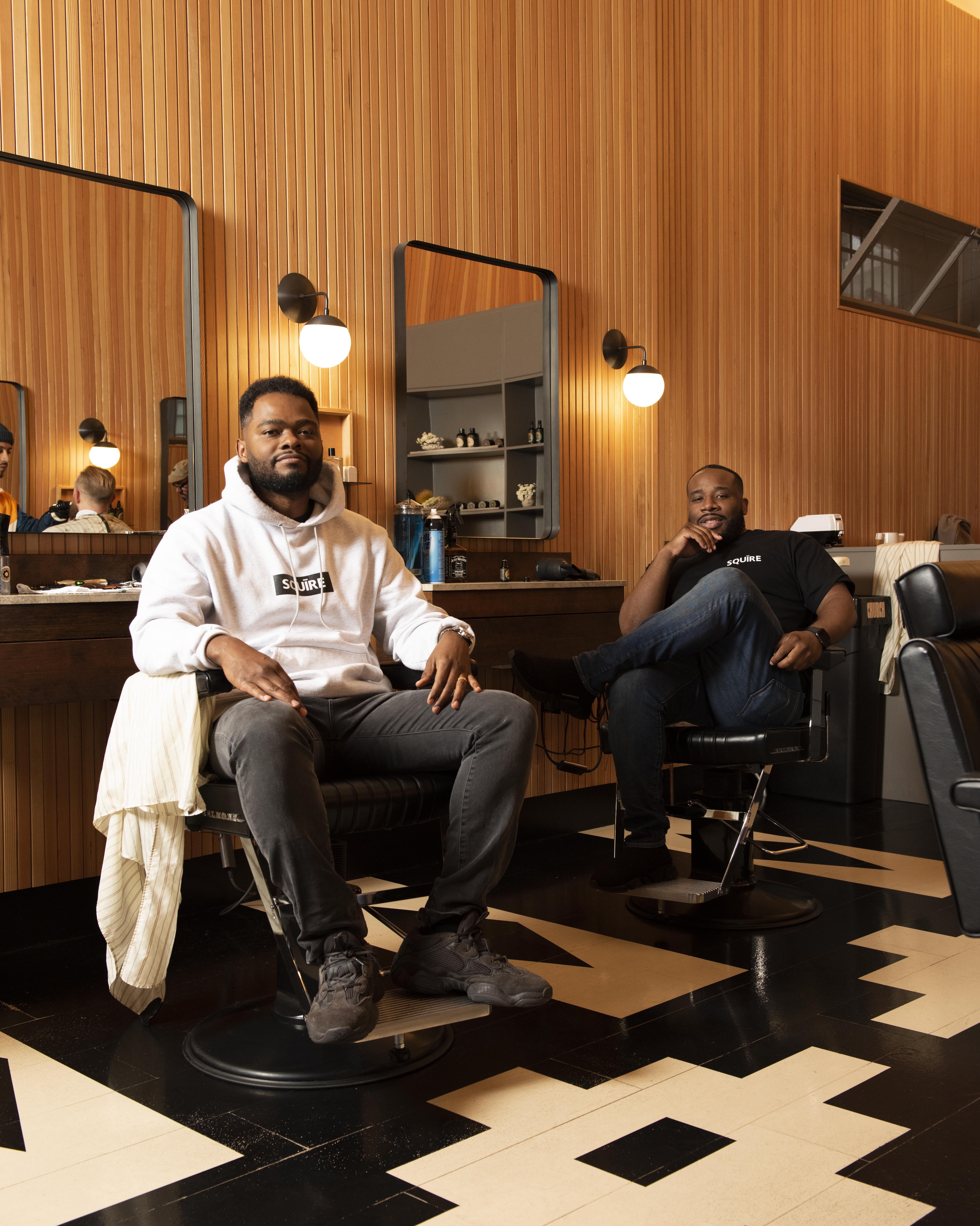 Two Black men, founders of the app Squire, sit in barber chairs in a barbershop setting. 