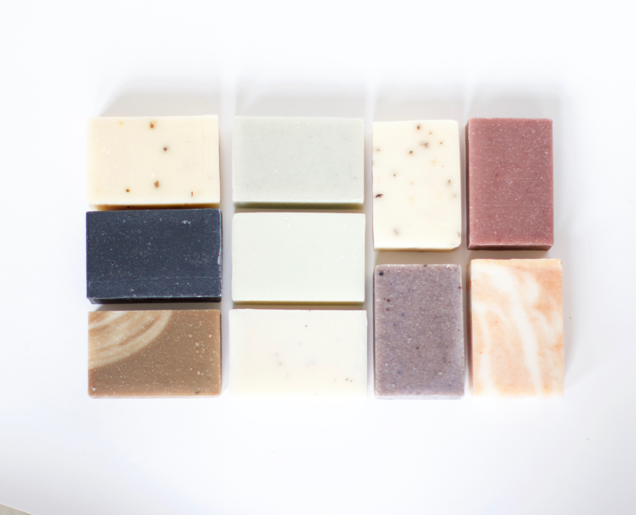 Ten different soap varieties, in a grid pattern, on a white background. Soaps vary in color from ivory to nearly-black, swirled peach, mauve and lavender.