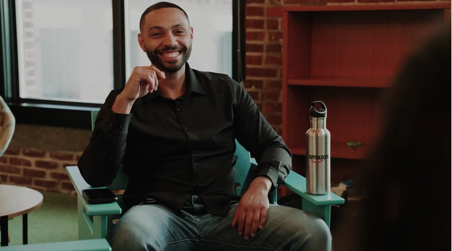 Smiling man in black shirt seated in teal chair with Amazon water bottle nearby