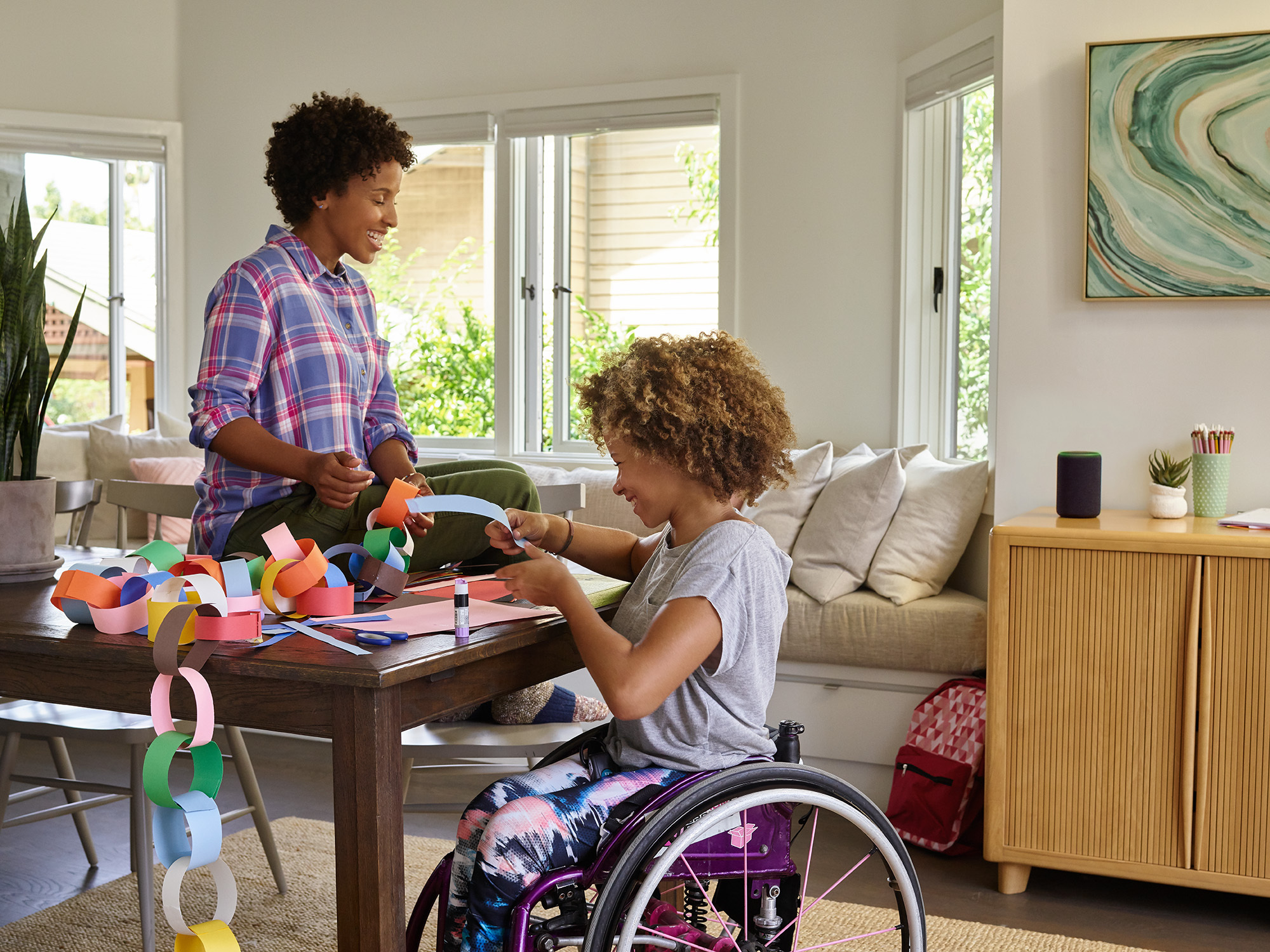 A mother sits on a dining room table as she and her daughter laugh, while making a paper chain. Behind them, Amazon Echo is on a console table. 