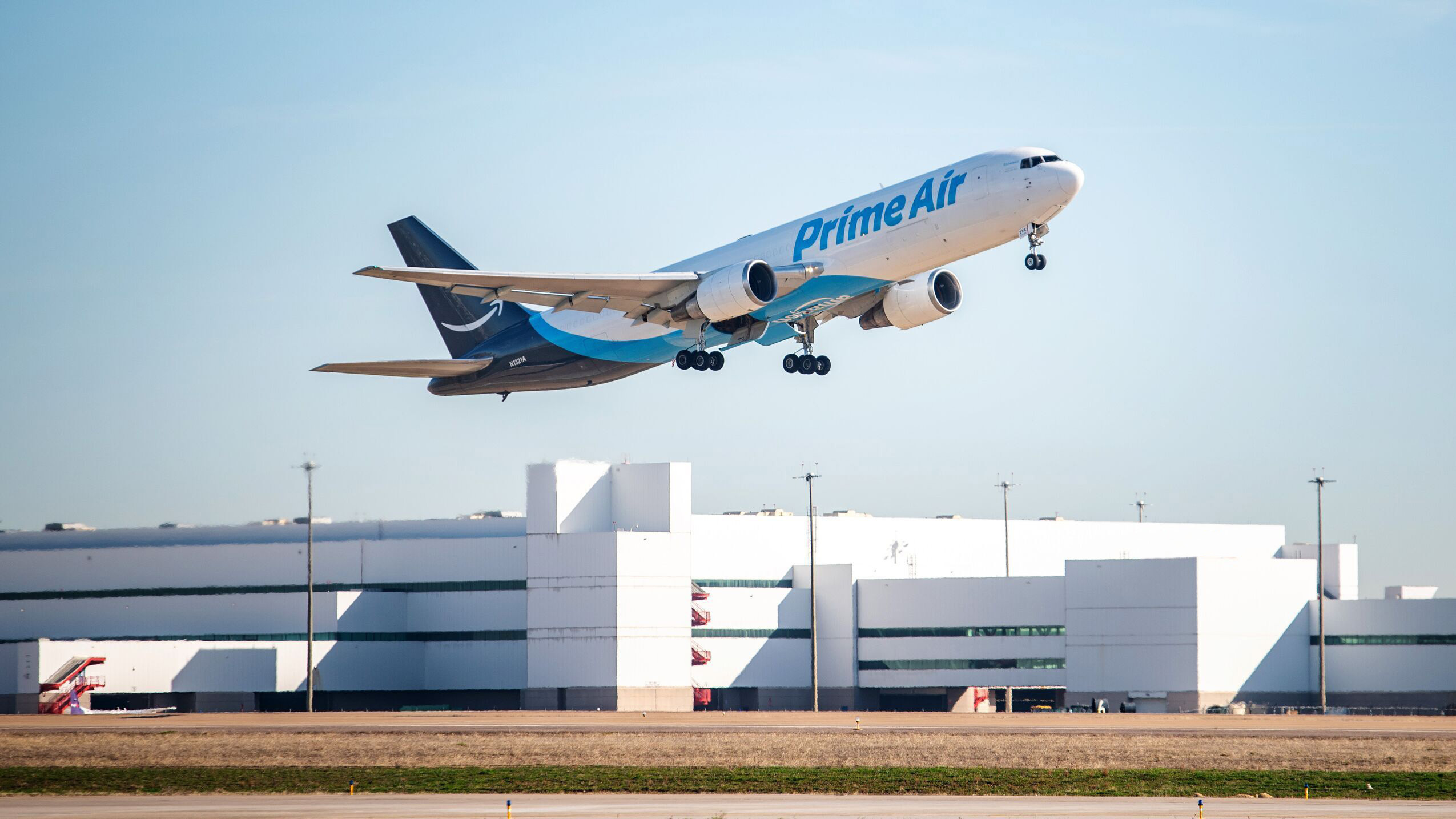 An image of an Amazon Air plane taking off and just leaving the ground from the runway. The plane is white with blue writing that says "Prime Air" and a white Amazon smile logo on the tail. There is a white building in the background.