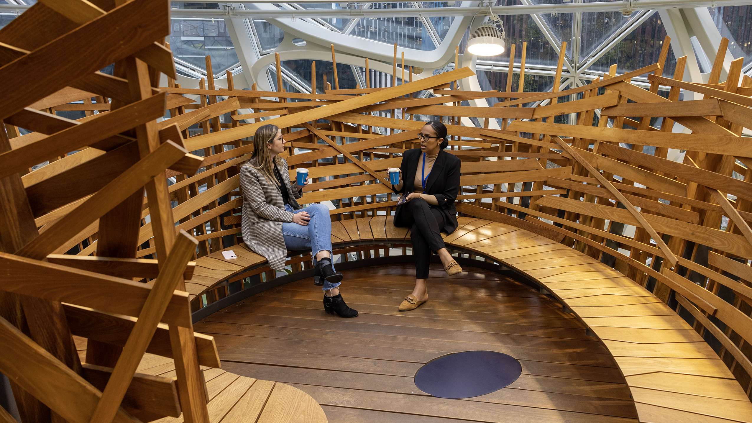 Two women sit inside a larger wooden structure drinking coffee and speaking.