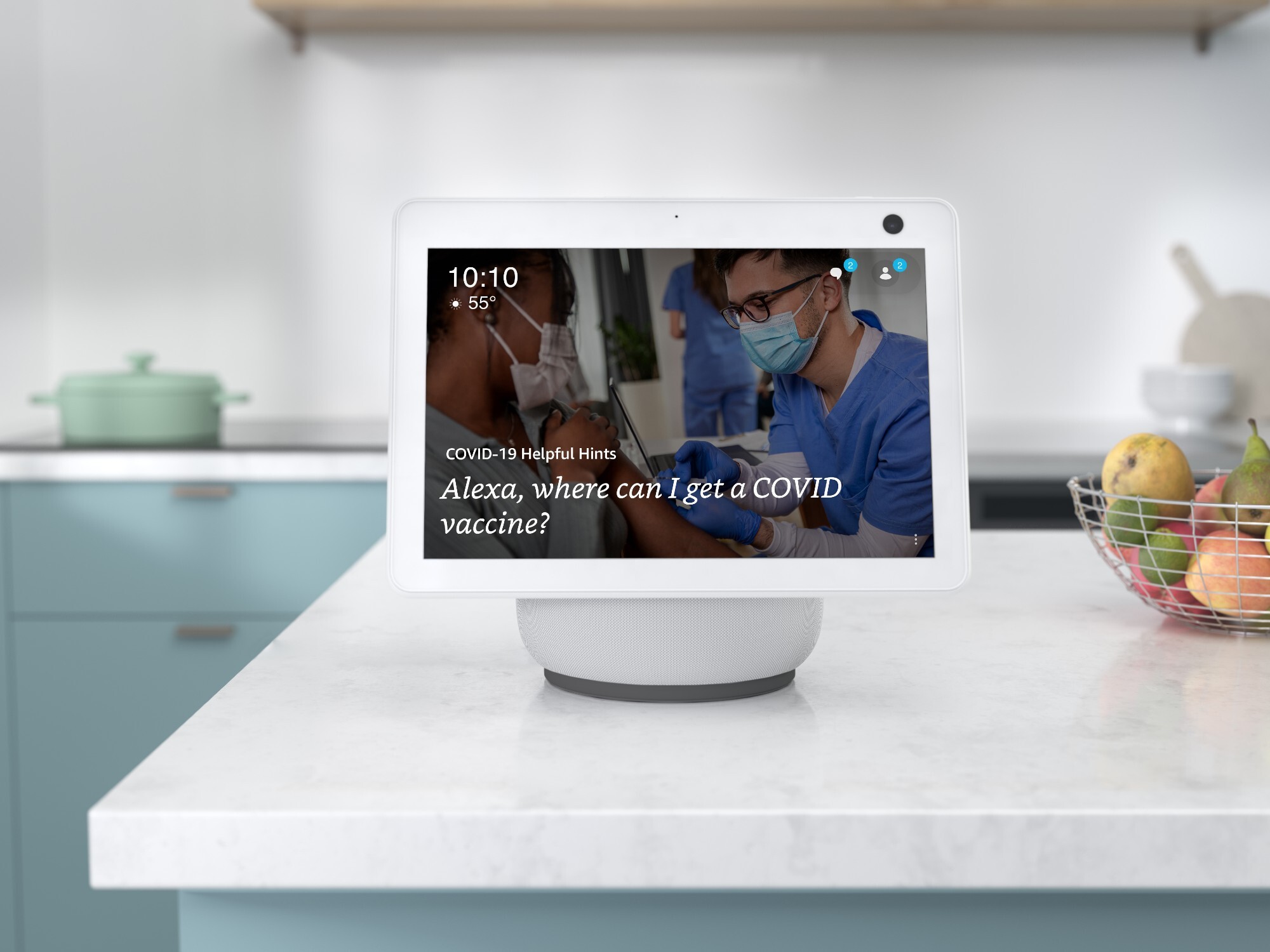 An Alexa device sits atop a white kitchen countertop and the screen reads "Alexa, where can I get a COVID-19 vaccine?" There is a wire basket filled with pears, limes, and apples next to the device. In the background there is a seafoam colored pot on the stove.