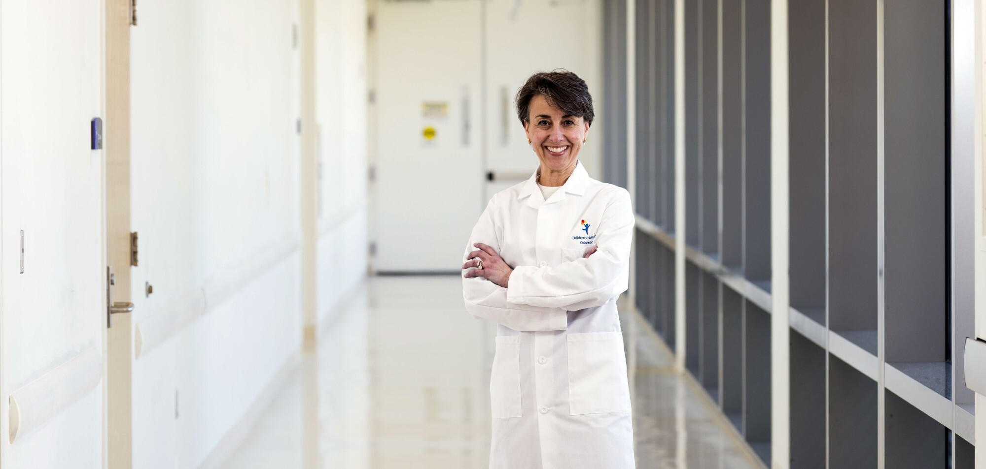 Childhood cancer researcher, Dr. Lia Gore, stands in the hallway of Children's Hospital Colorado smiling for the photo in her white doctors' coat. 