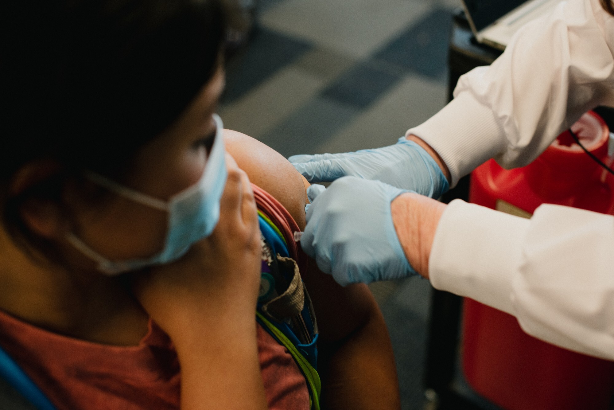 A woman wearing a mask holds up her sleeve as she receives the COVID-19 vaccine from a nurse wearing a white jacket and blue gloves.