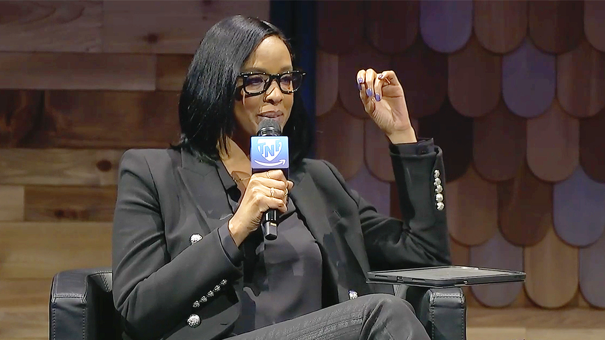 An image of Amina Hussein sitting on a panel discussion and holding a microphone with a "TNF" logo on it.  