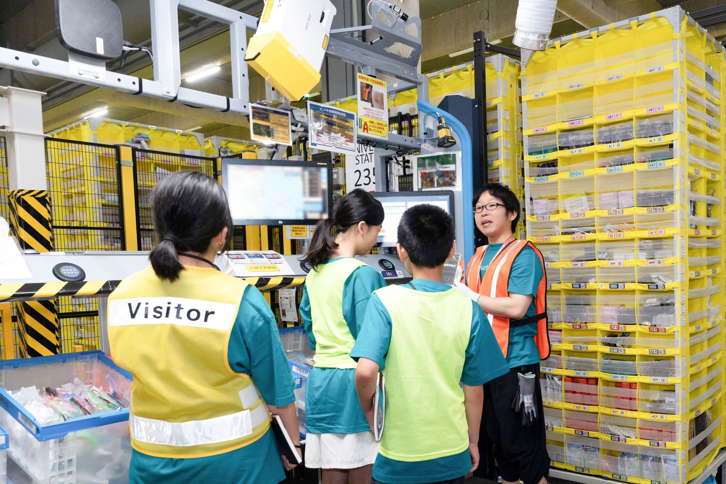 黄色の棚の前で、3人の子どもたちに話しかけているオレンジ色の安全ベストを着ている男性