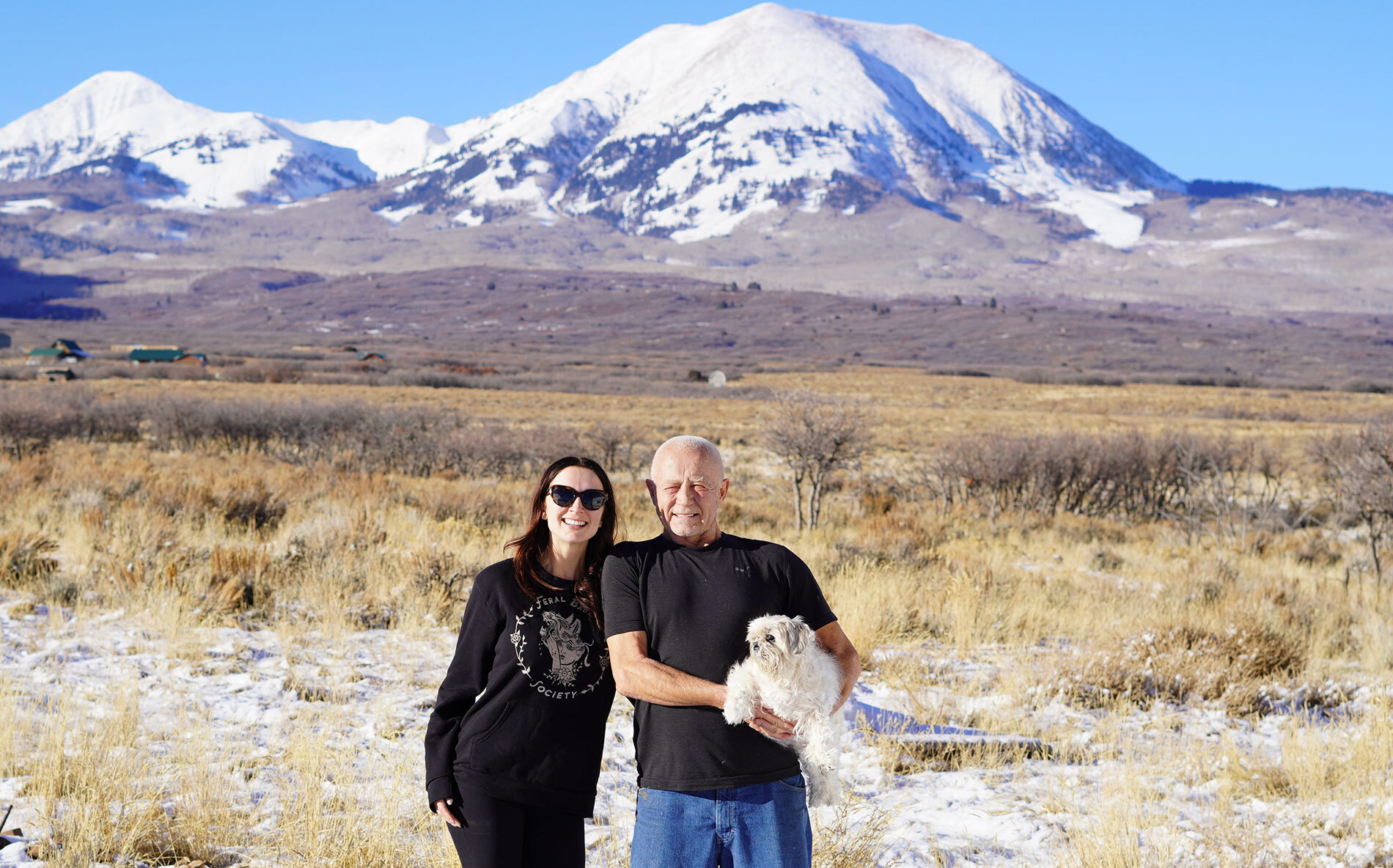 An image of a man and his daughter smiling for a photo. The man is holding a small white dog in his arms. There is a landscape in the background with a large, snow-capped mountain.