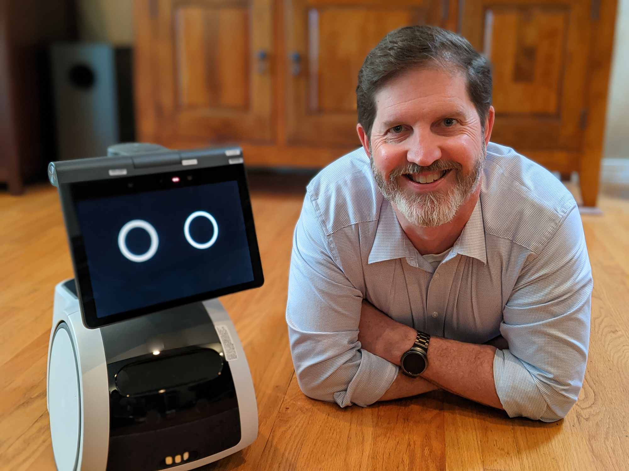 A man lays on the floor and smiles as the camera, next to an Amazon Astro house robot, in a kitchen.