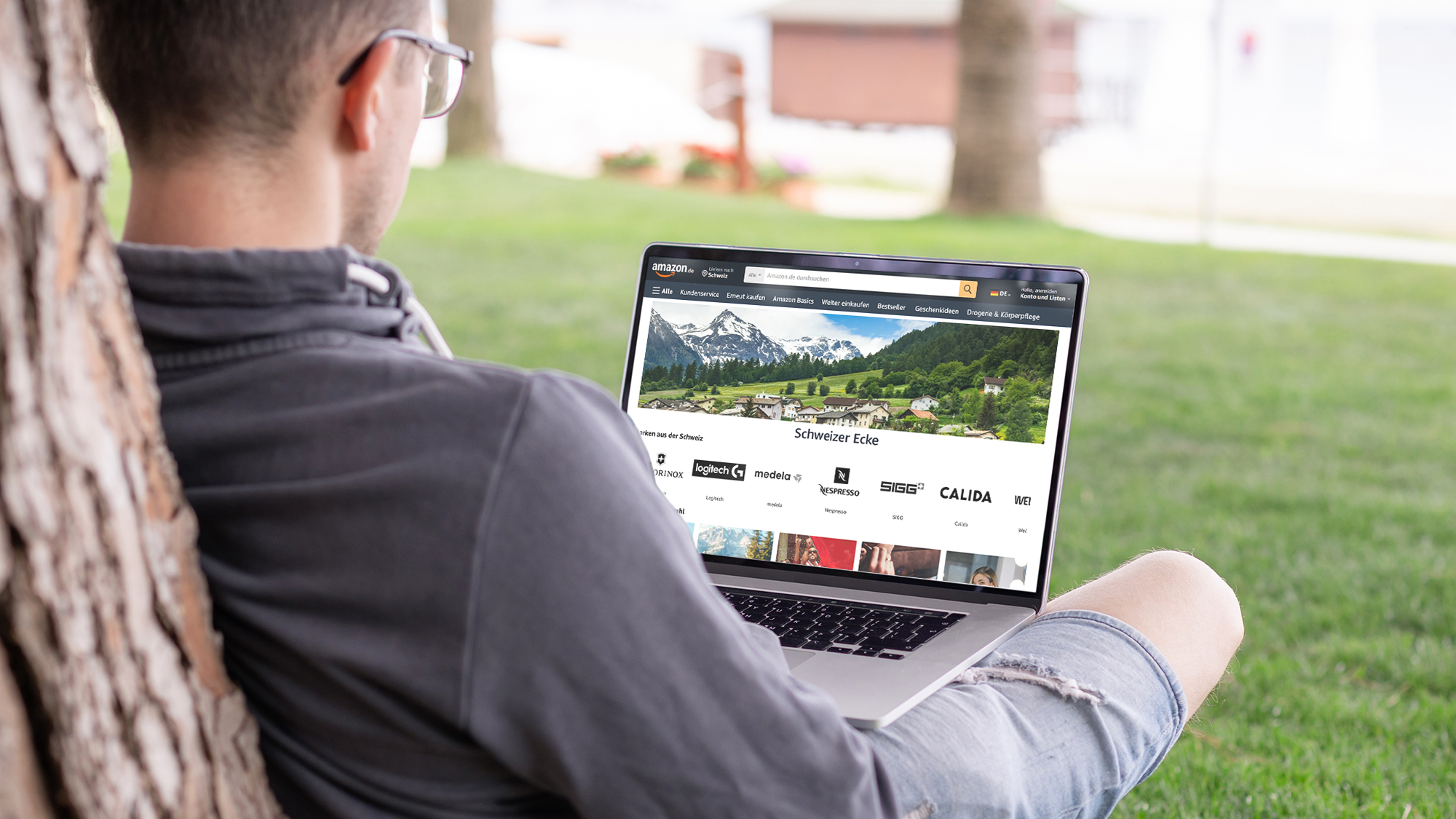 Aus der Schweiz bei Amazon bestellen: Das Foto zeigt einen Mann mit kurzen Haaren und Brille von hinten, er sitzt auf einer Wiese, angelehnt an einen Baum. Auf seinem Schoß ein Laptop, der eine Seite auf amazon.de zeigt, die mit "Schweizer Ecke" betitelt ist.
