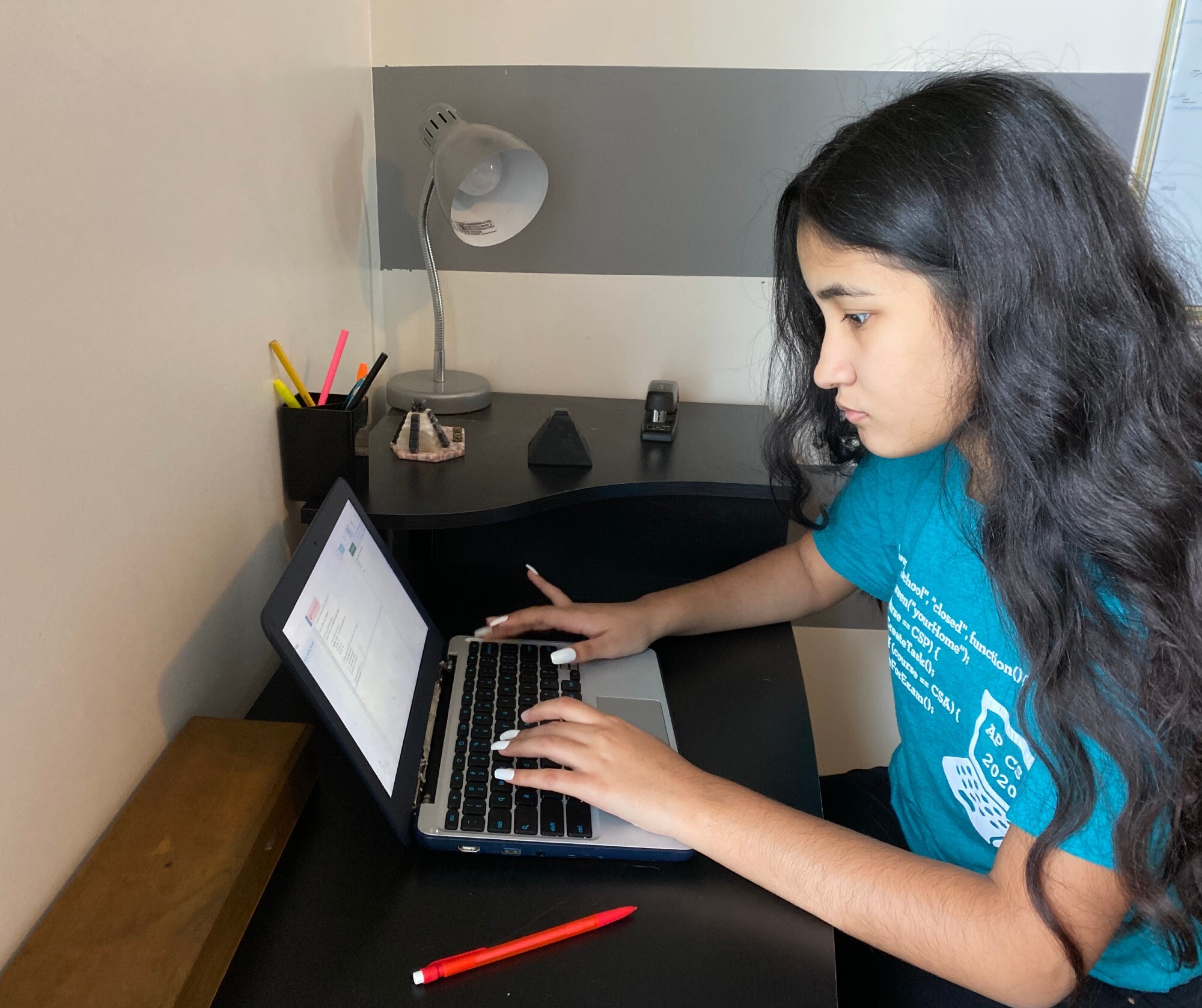 A young woman types on a laptop at a desk in her home. She wears a blue t-shirt from a coding program.