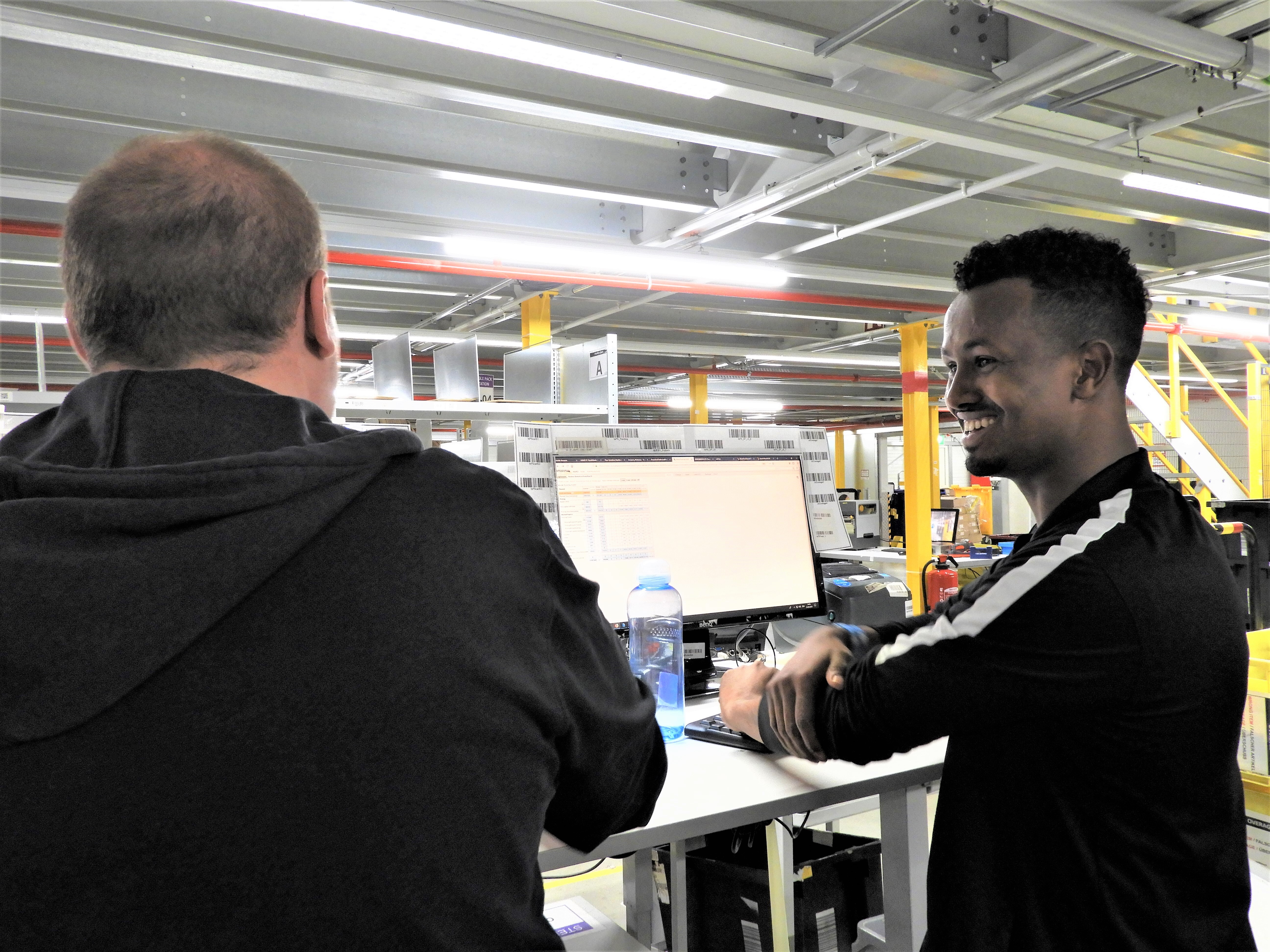 2 Mitarbeiter vor einem Laptop. Den linken Mitarbeiter sieht man von hinten, der rechte blickt lächeln zu ihm. Beide tragen dunkle Kleidung, nur die Oberkörper sind zu sehen.