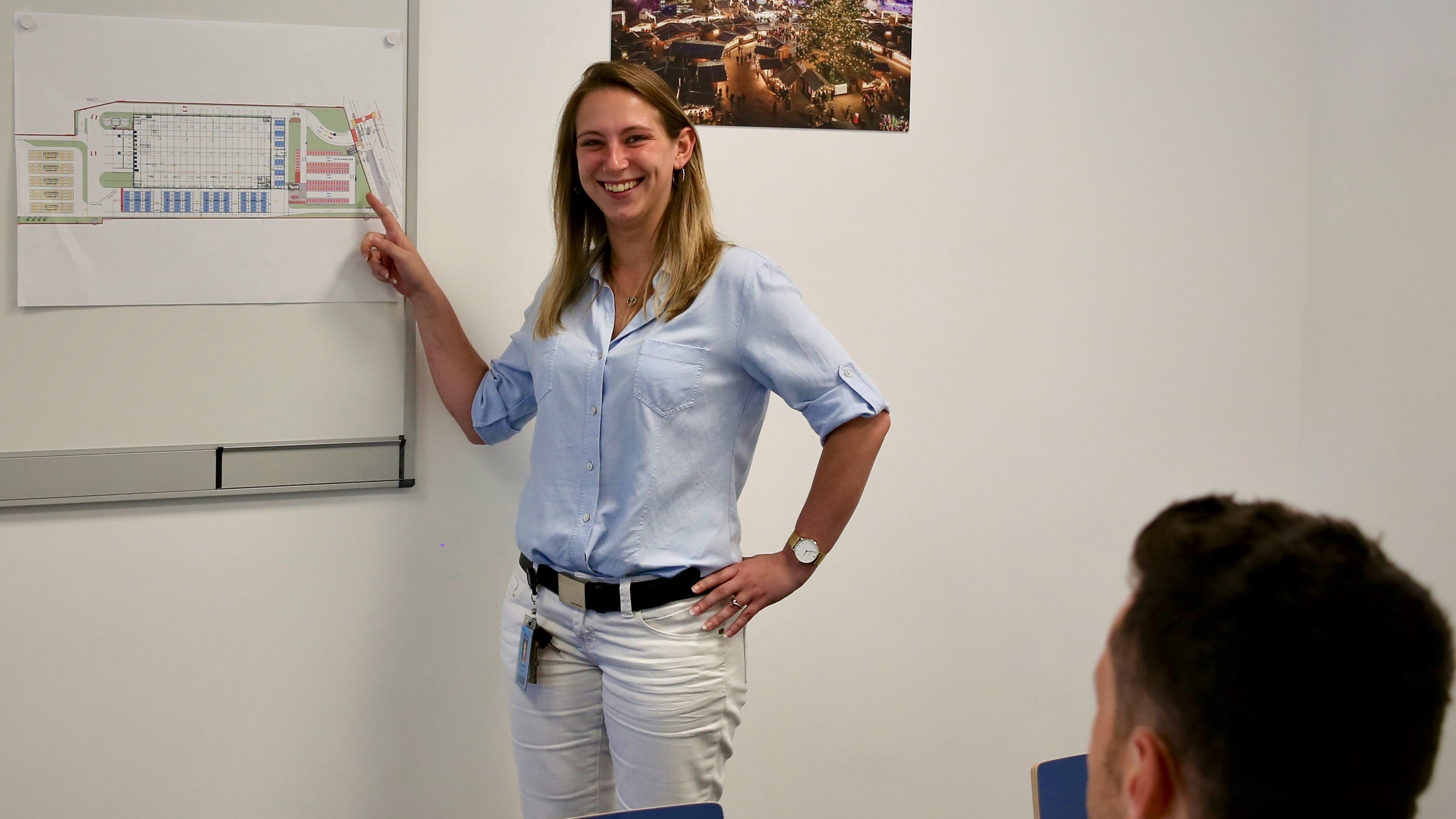 Mitarbeiterin mit halblangen blonden Haaren, blauer Bluse, weißen Jeans, vor dem Flipchart