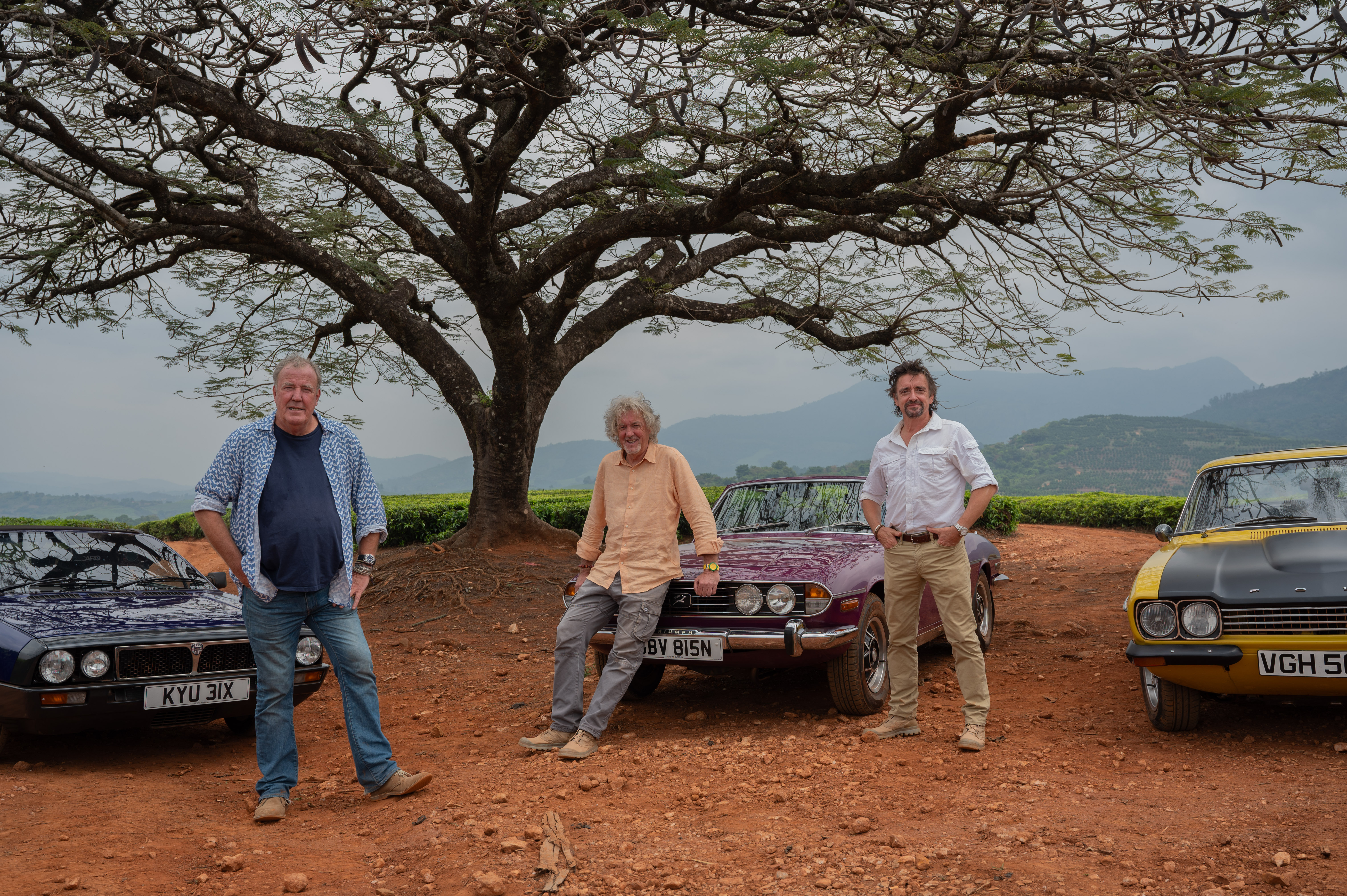 three men stand next to three vintage cars in a rural area