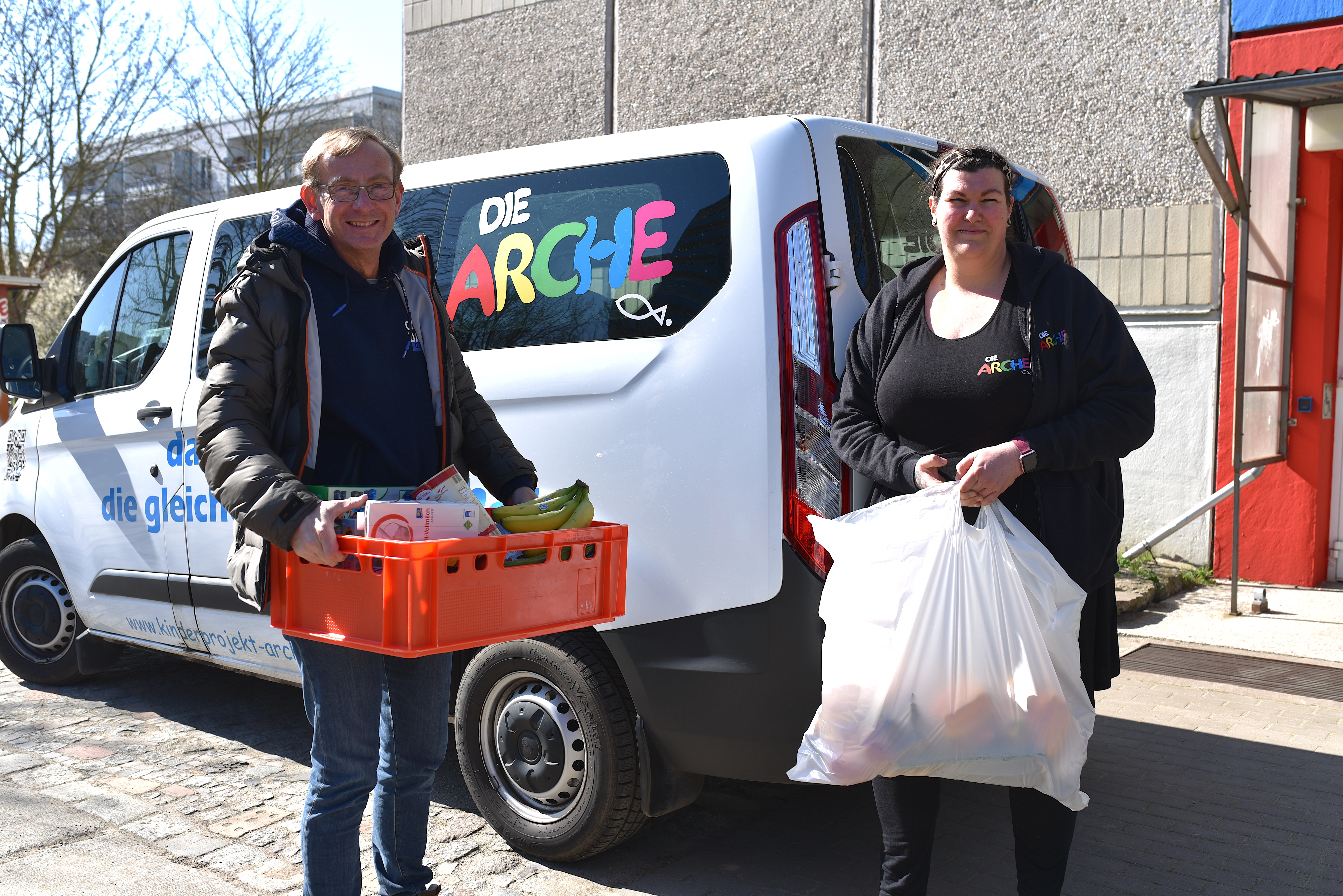 2 Mitarbeiter der Arche mit Lebensmitteln vor einem Lieferwagen mit Aufdruch "Die Arche".